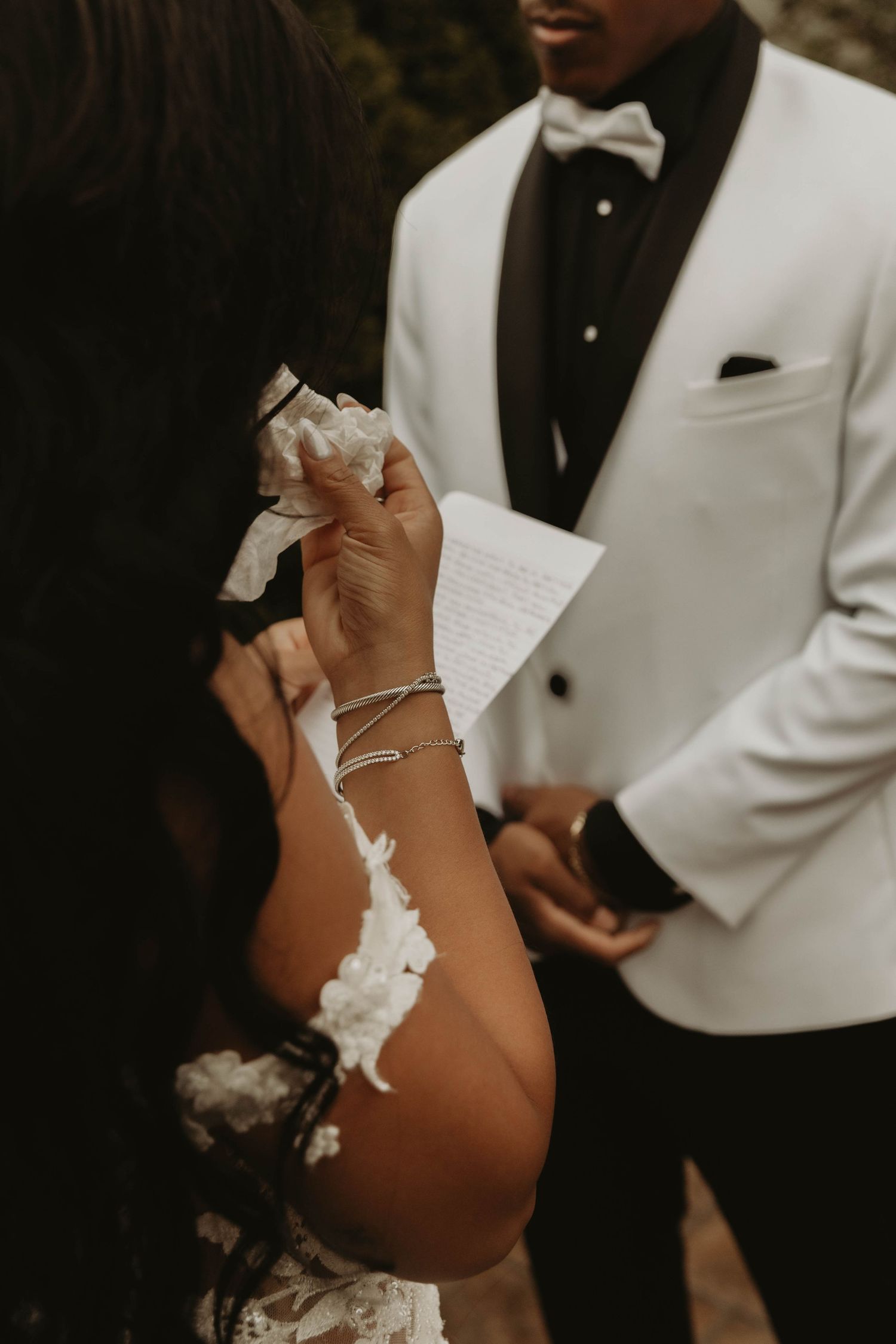 Emotional bride wiping tears during wedding vow exchange as groom in white tuxedo reads from paper.