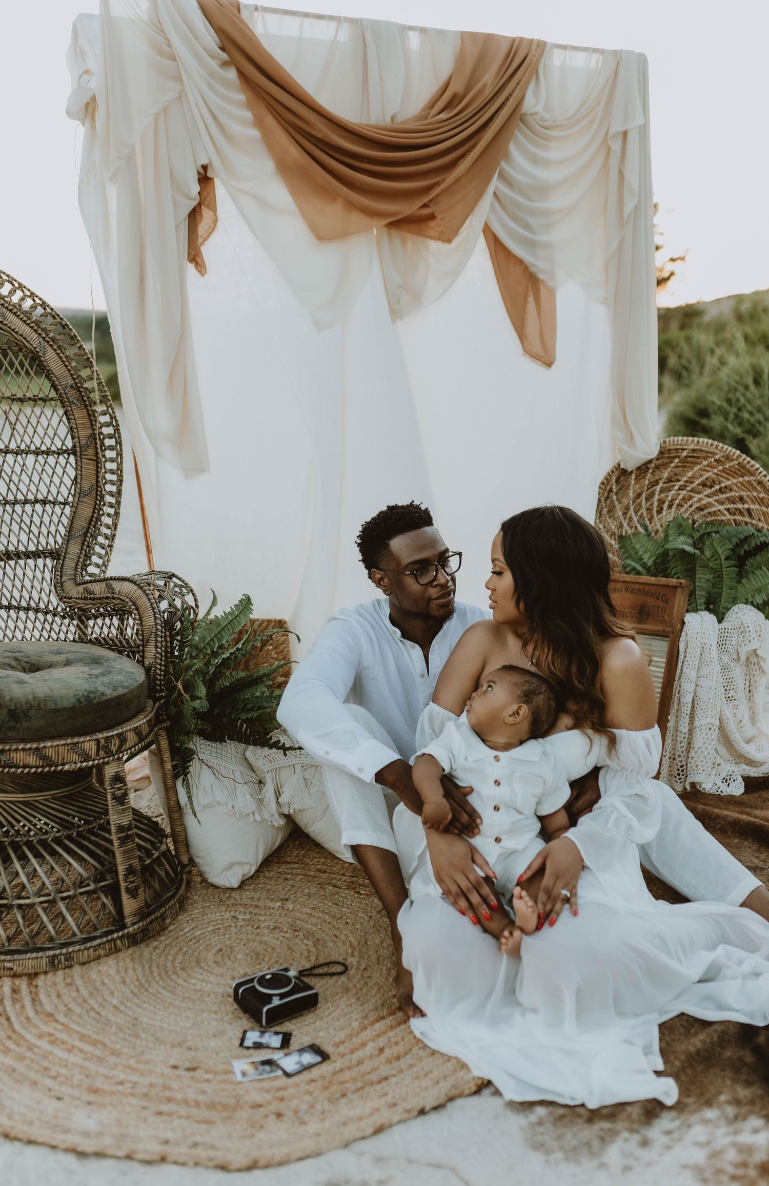 A couple in white outfits sits on a boho rug with their newborn baby, surrounded by wicker chairs and draped fabric.