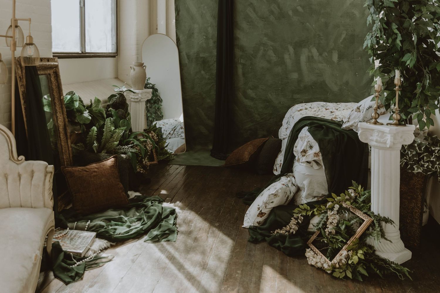 A rustic boho studio with green walls, scattered foliage, pillows, and vintage decor bathed in natural sunlight.