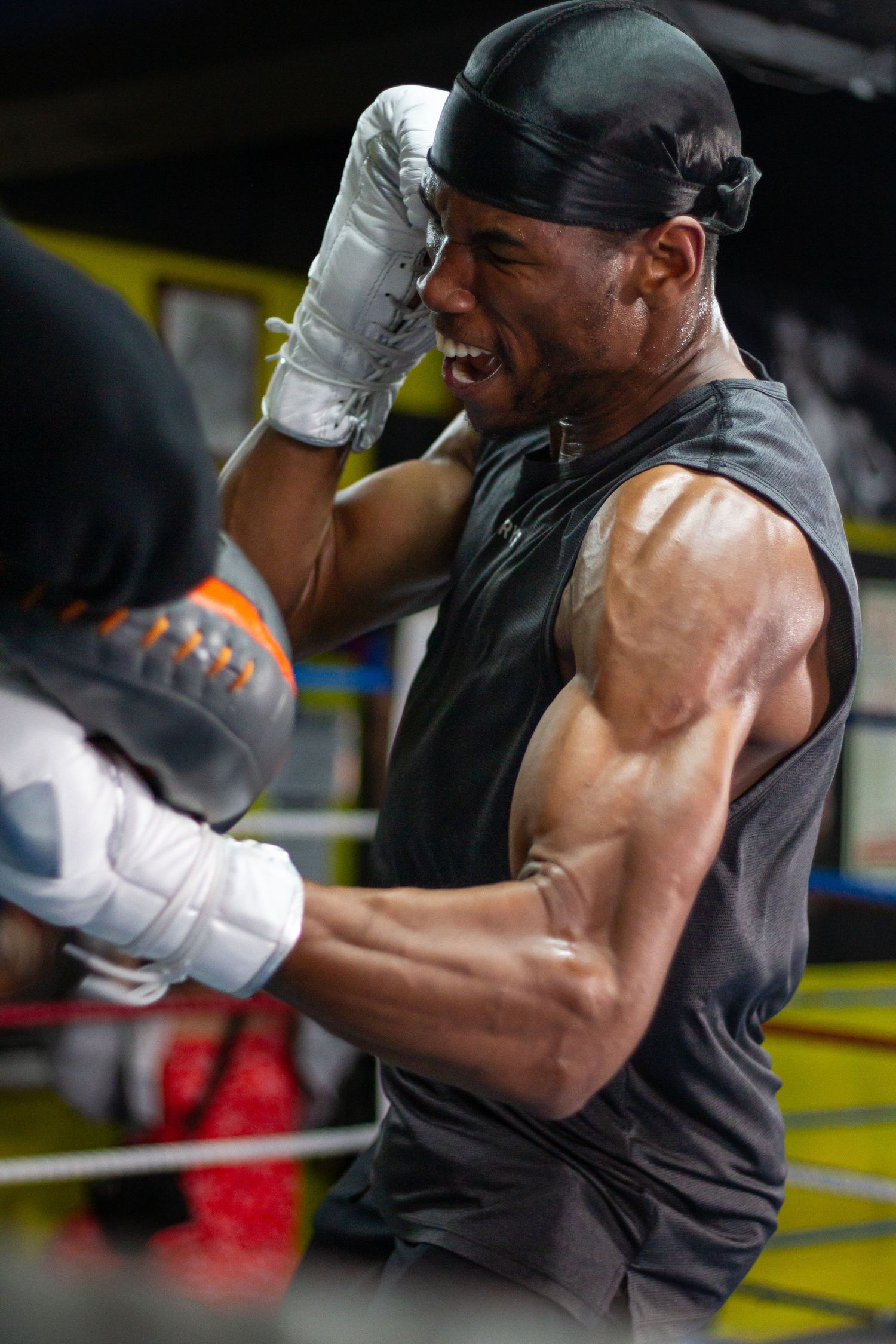 Muscular boxer sparring in gym wearing white gloves and black sleeveless shirt during intense training session.