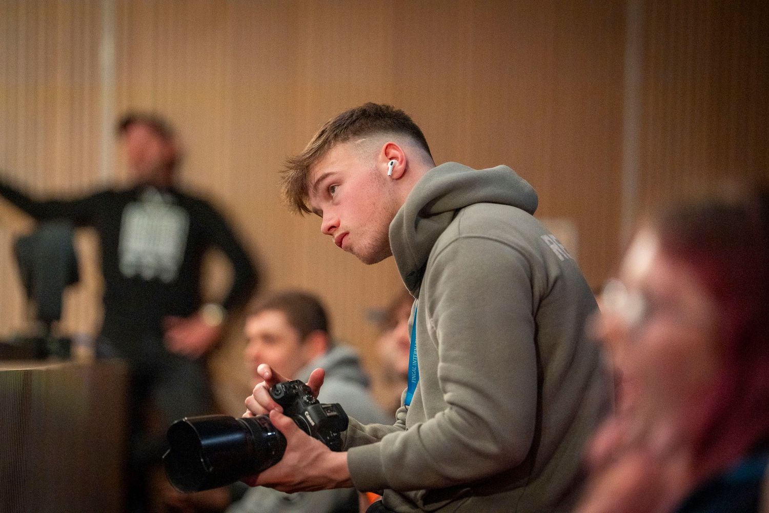 Young man in grey hoodie holding a camera in a modern conference room setting.