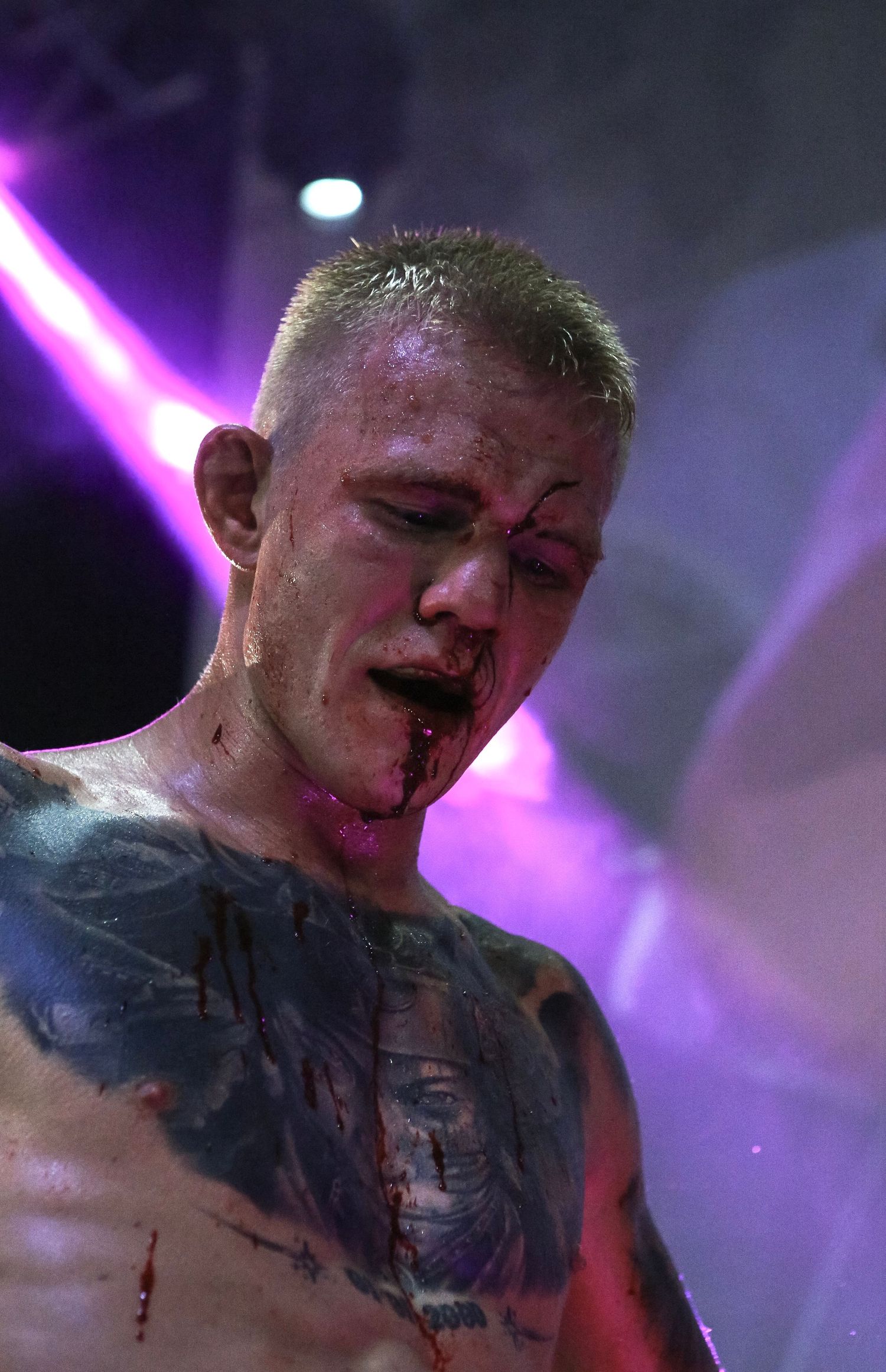 Bloodied male MMA fighter with tattoos under purple stage lights after intense combat sports match.