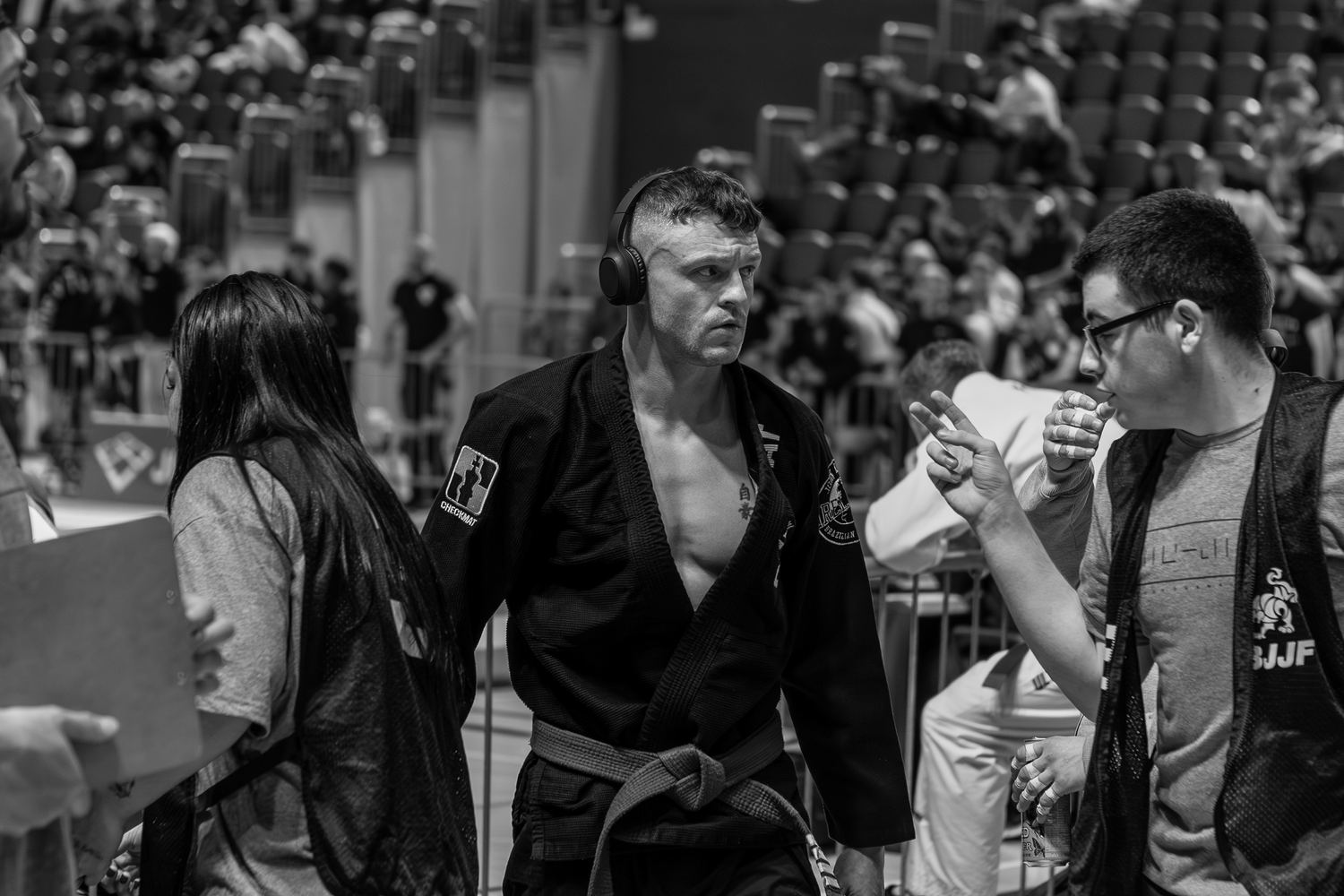 Male BJJ competitor in black gi walks confidently at a jiu-jitsu tournament surrounded by officials.