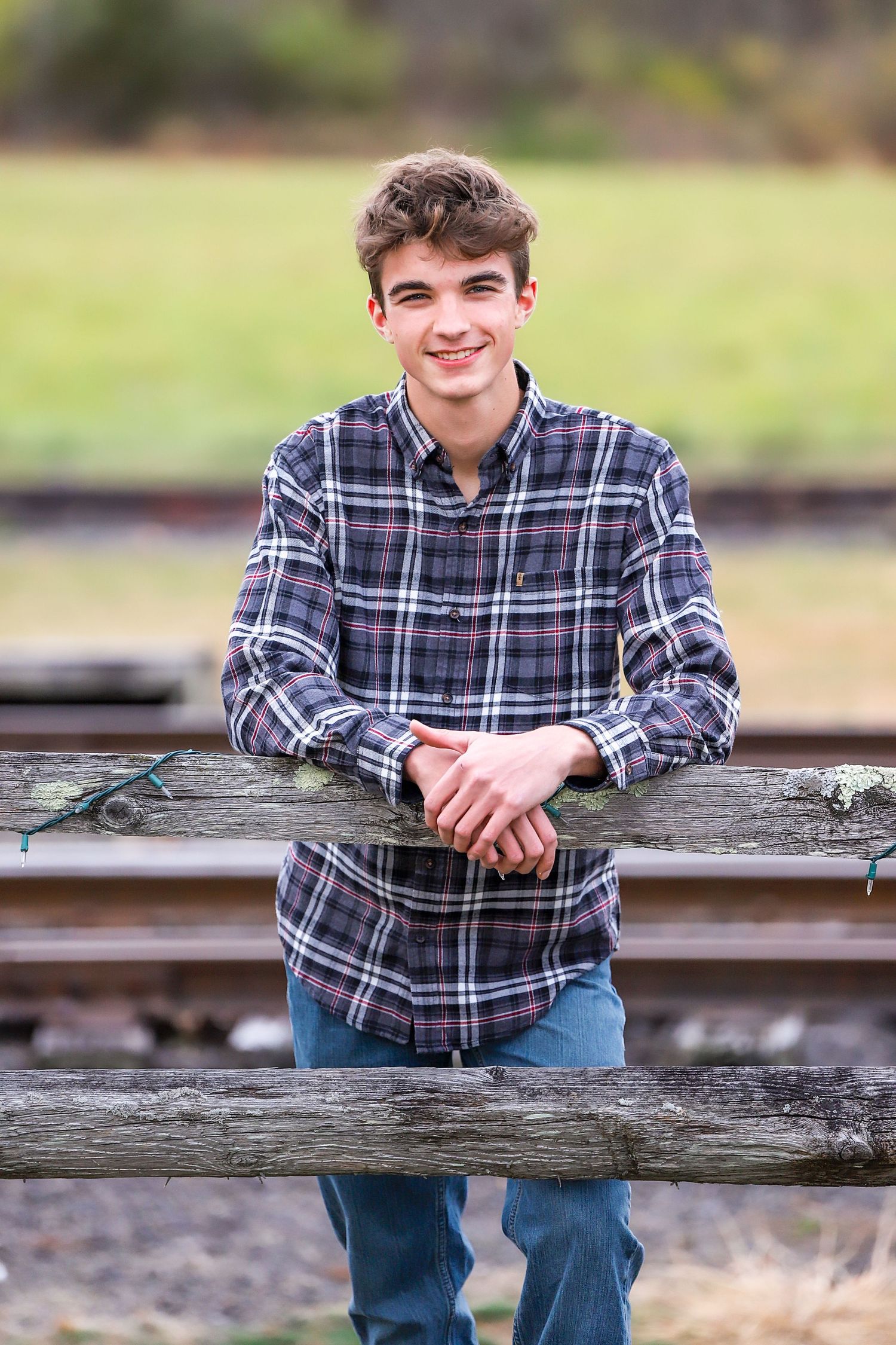 Young person in plaid shirt leaning casually against wooden fence outdoors.