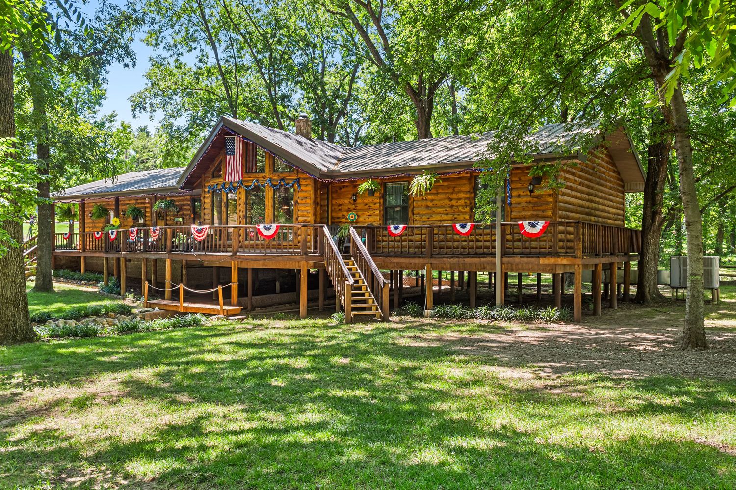 Elevated wooden cabin with wrap-around deck nestled among green trees and landscaped yard.