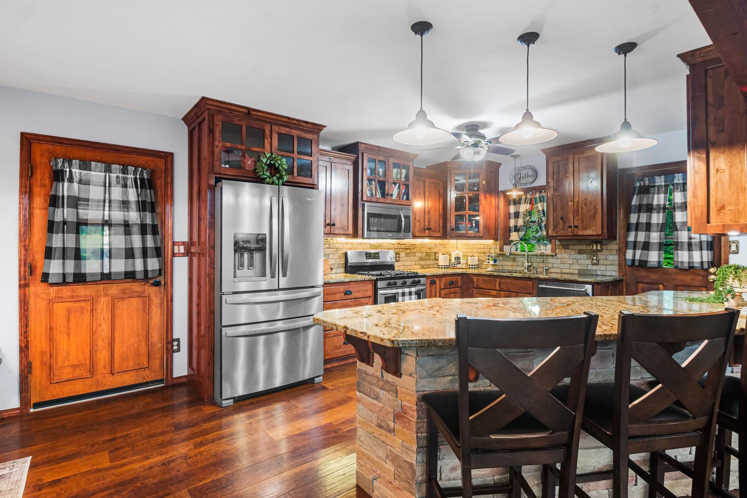 Country kitchen with rich wood cabinets, stainless appliances, and buffalo check curtains.