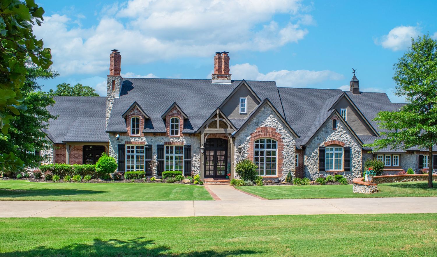 Luxurious stone and brick mansion with dormers and multiple chimneys set on a manicured lawn under blue skies.