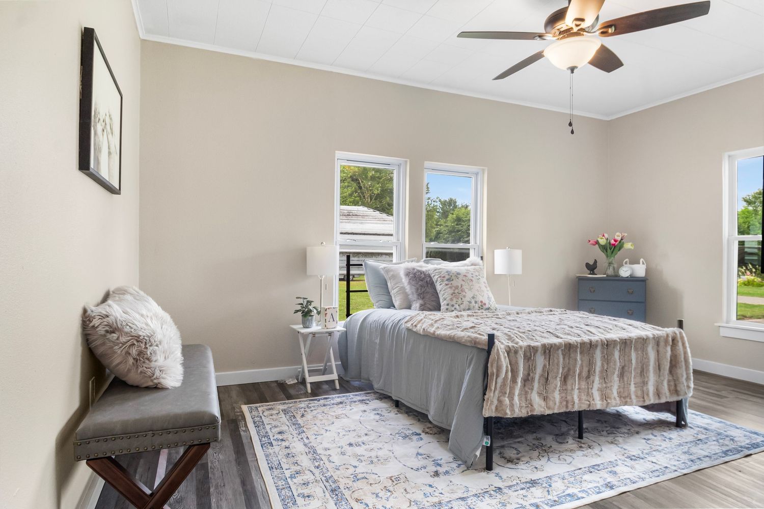 Bright bedroom with ceiling fan, gray bedding, and multiple windows offering natural light.