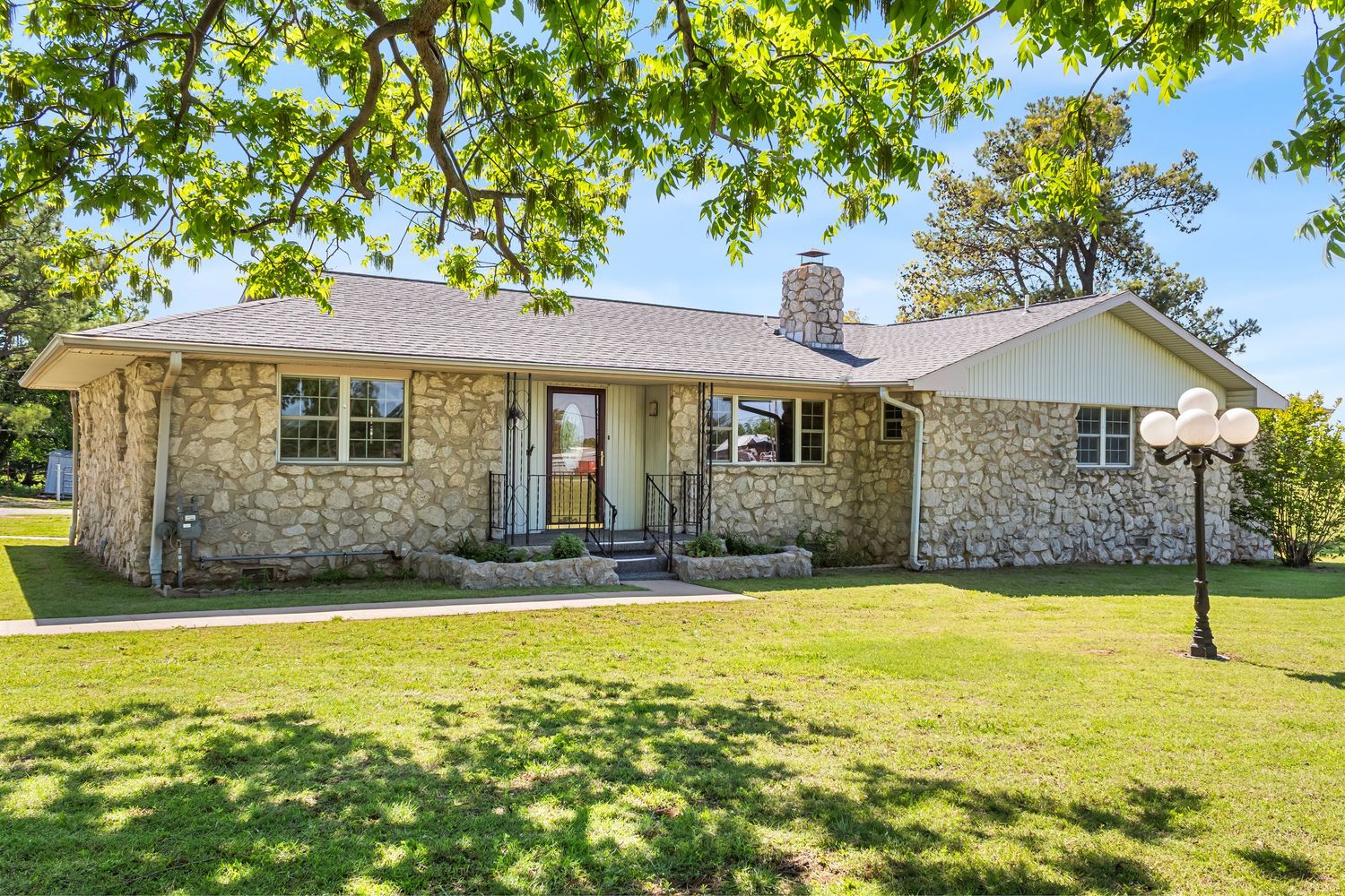 Stone and brick ranch house with large front yard and mature trees in rural setting.