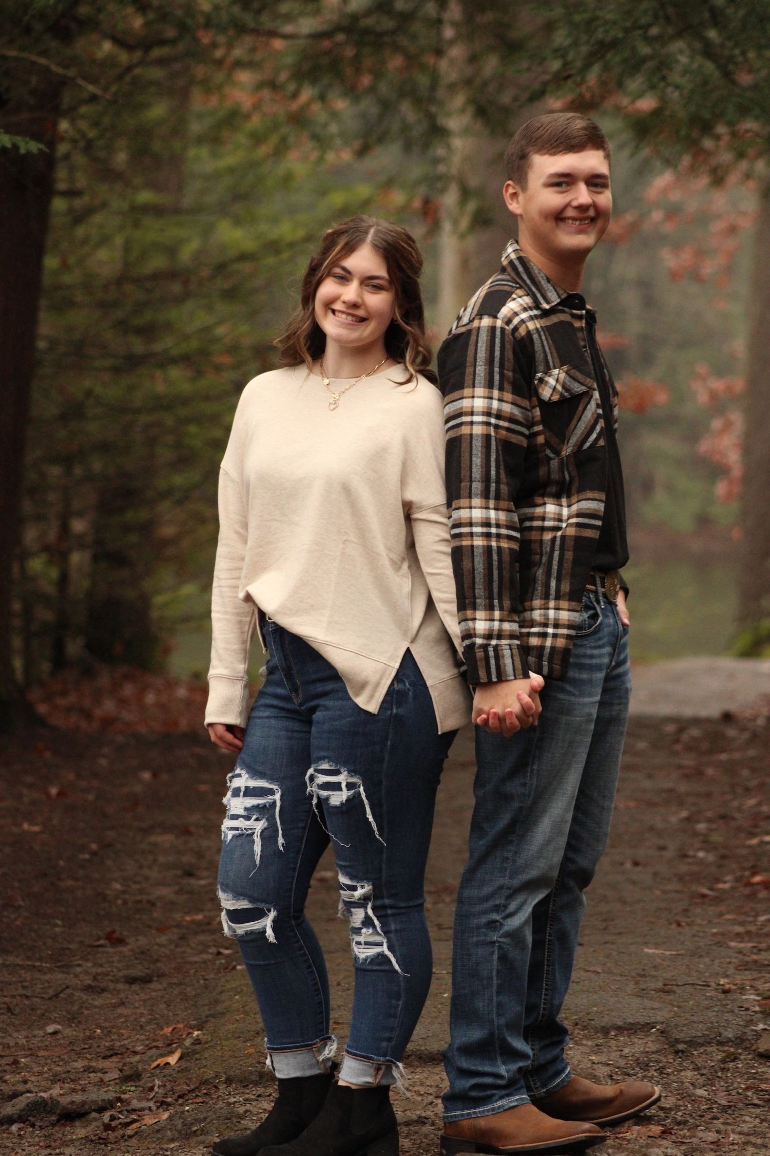 A couple in casual fall attire stands together on a leaf-covered forest path during autumn.