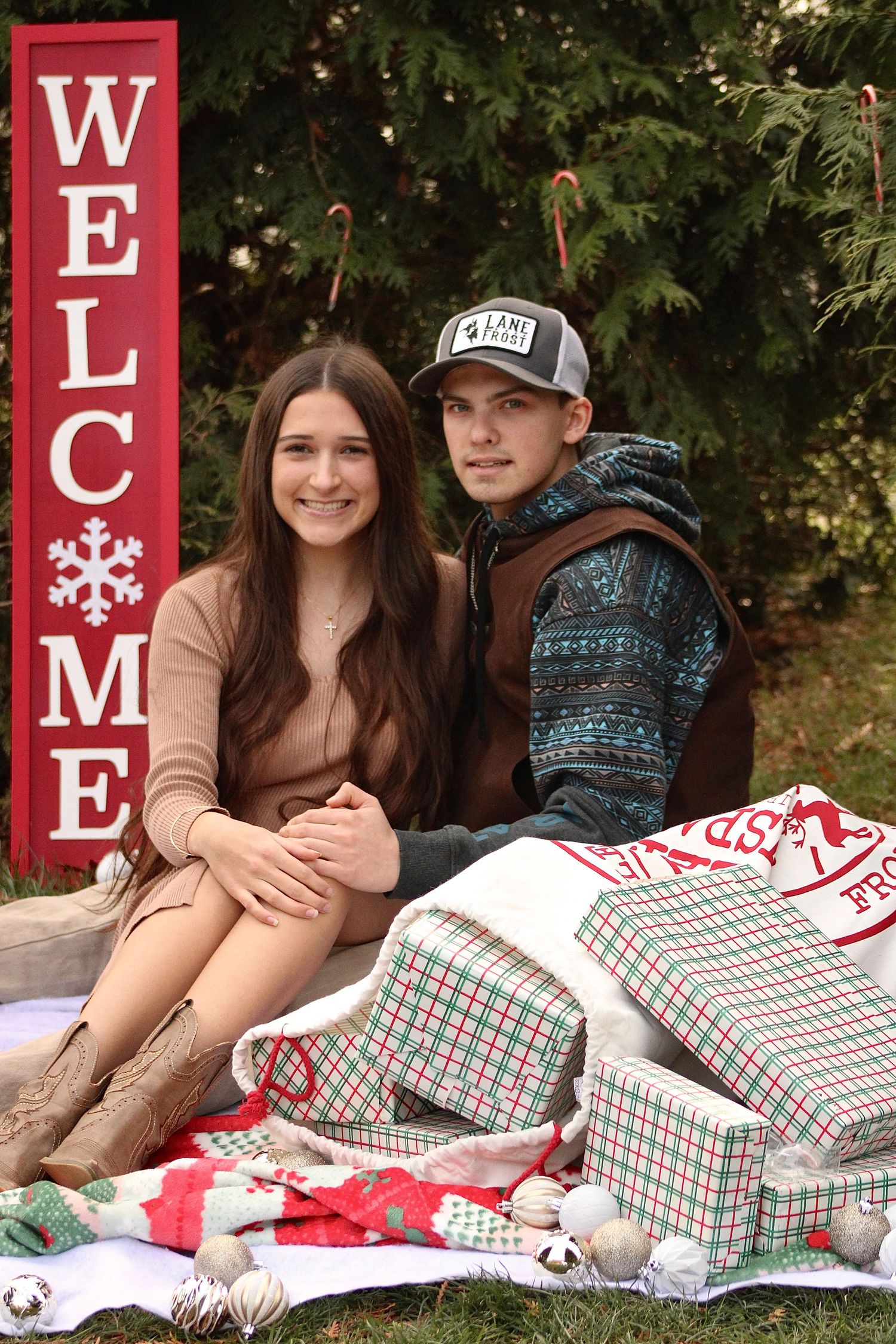A couple sits on a white blanket with red and green plaid pillows next to holiday welcome signs.