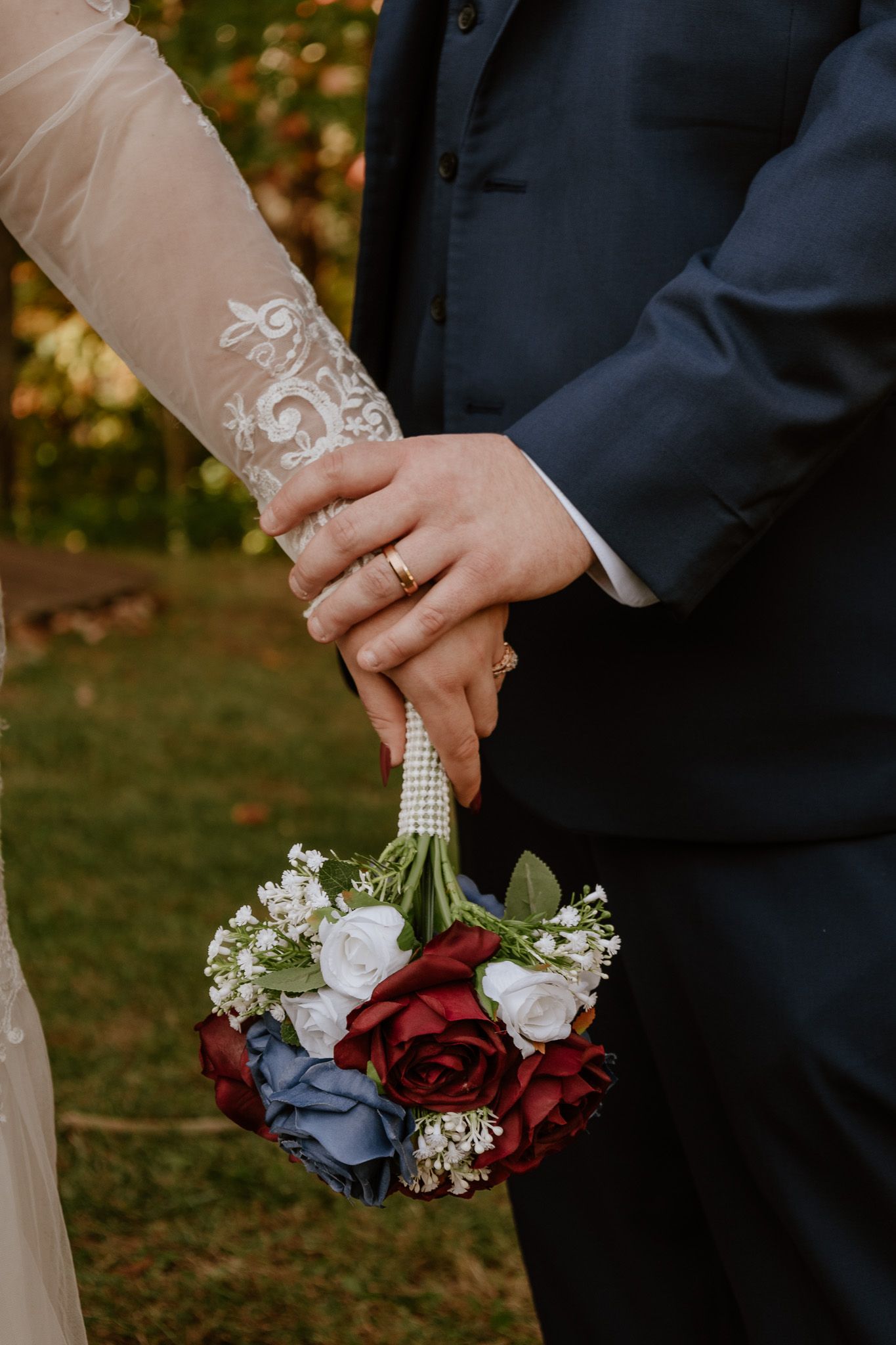 Close-up of wedding couple's hands holding a bouquet with red, blue and white roses wrapped in pearl details.