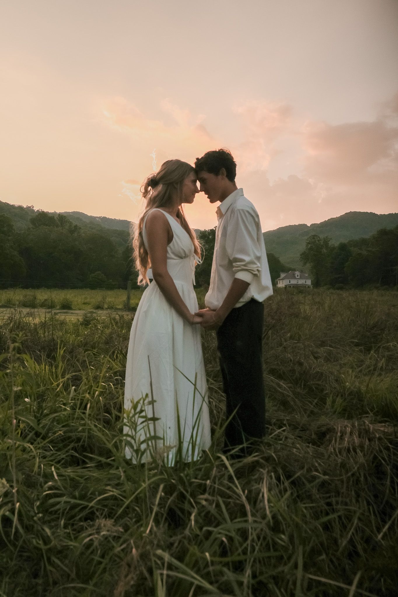 A couple shares an intimate moment at sunset in a field with mountains in the background.
