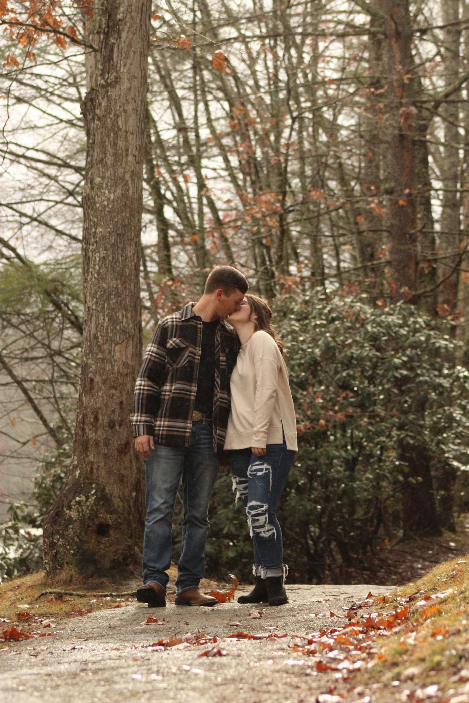 A couple shares a tender moment together in a snowy winter forest setting with fallen autumn leaves on the ground.