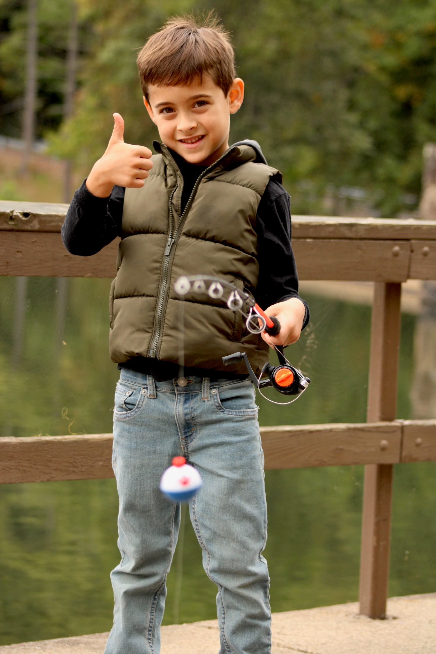 A person wearing jeans and a puffy vest gives a thumbs up while fishing at a wooden railing by a lake.