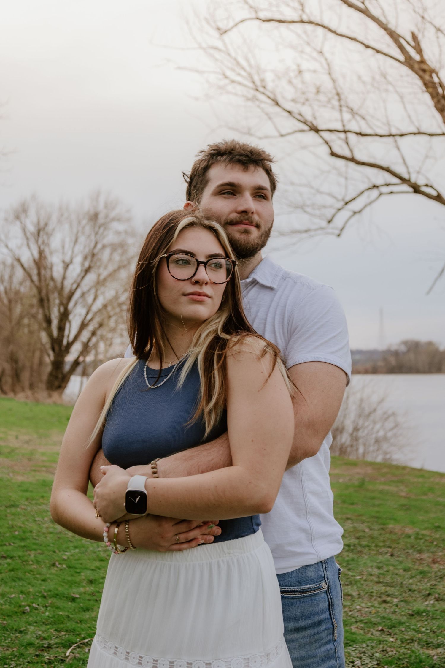 A couple embraces outdoors on a grassy hill during a romantic photoshoot near bare winter trees.