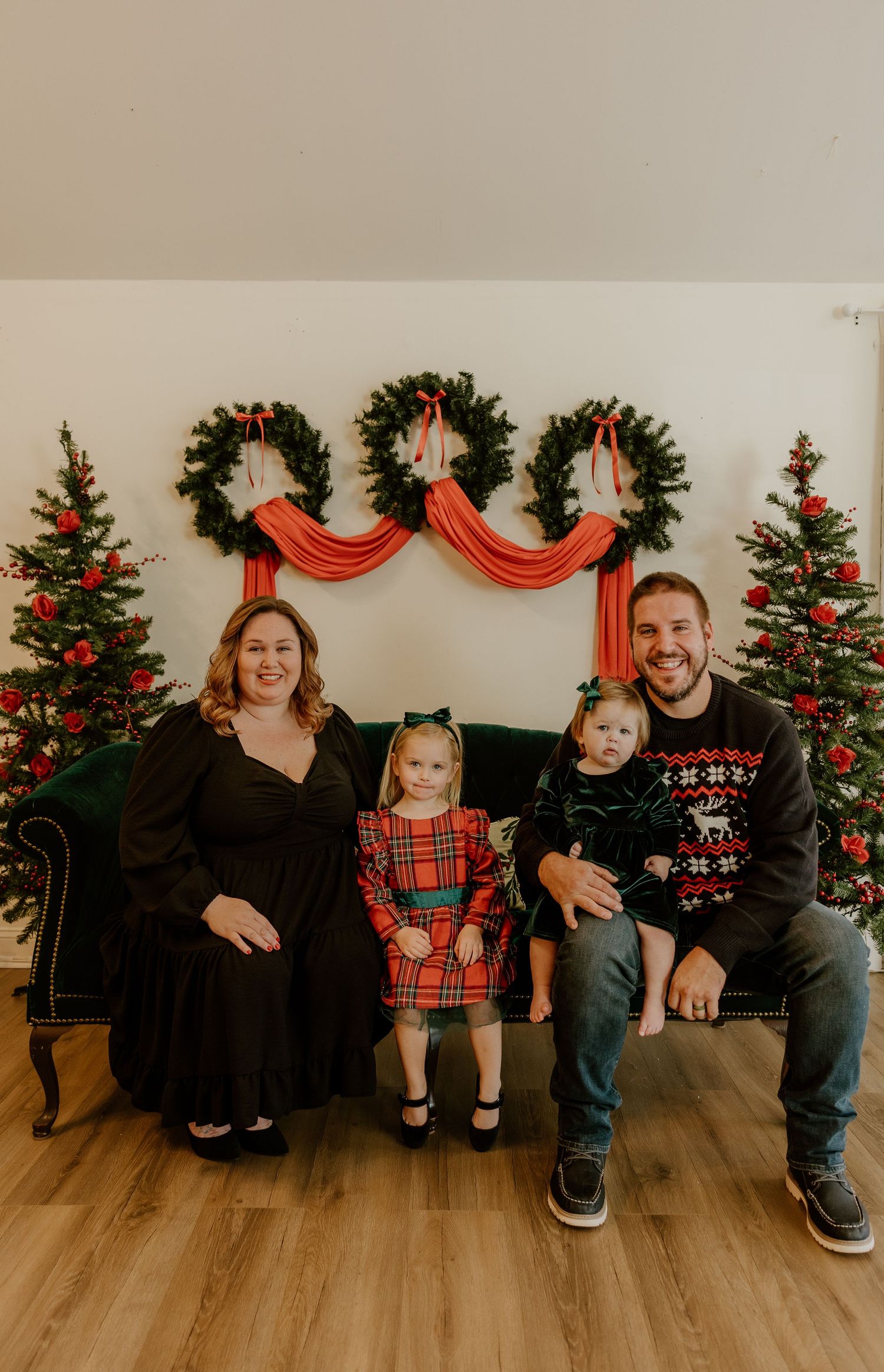 A family poses for a Christmas photo in front of three wreaths with red draping and decorated Christmas trees.