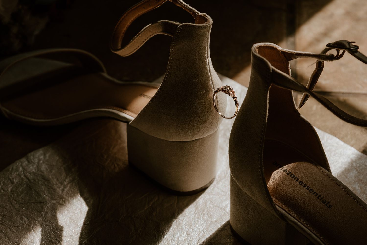 Elegant beige suede ankle strap heels casting dramatic shadows in moody lighting. Wedding details of ring and heels.