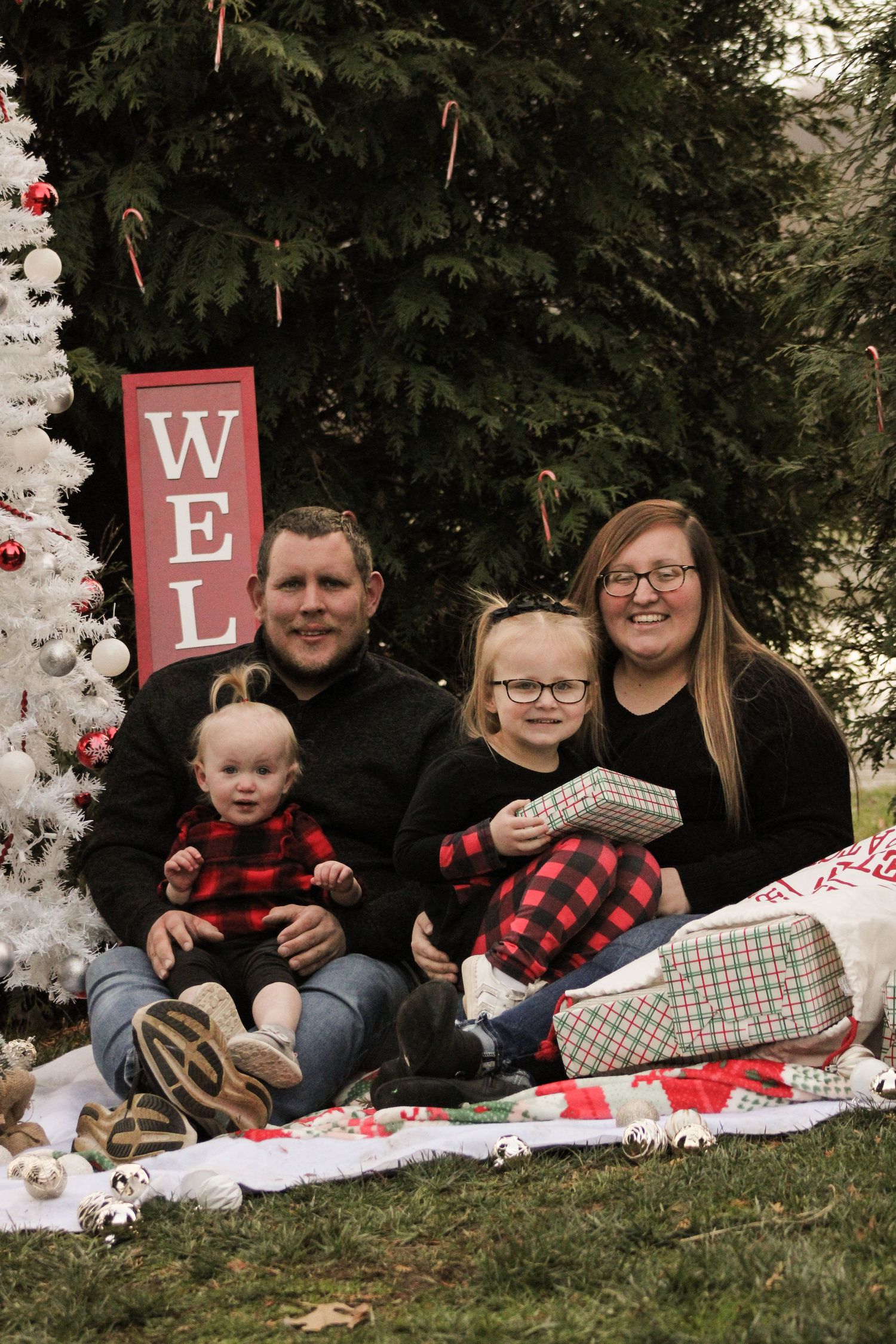Family wearing buffalo plaid sits together on white blanket for holiday photos with festive decorations.
