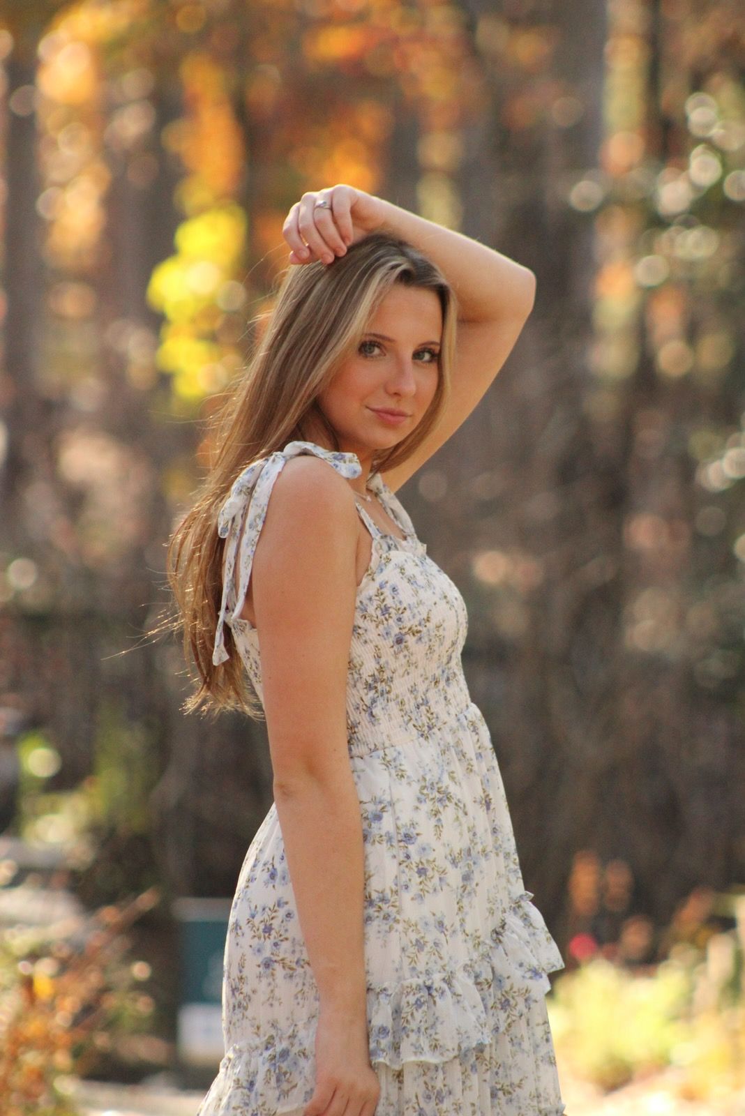 A person in a floral sundress poses playfully in a sun-dappled forest setting with golden light filtering through trees.