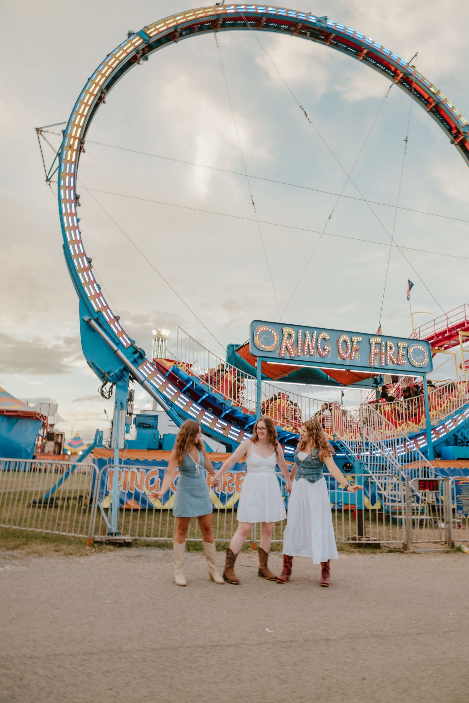 People walking past a large Ring of Fire carnival ride at a fairground with dramatic clouds in the sky.