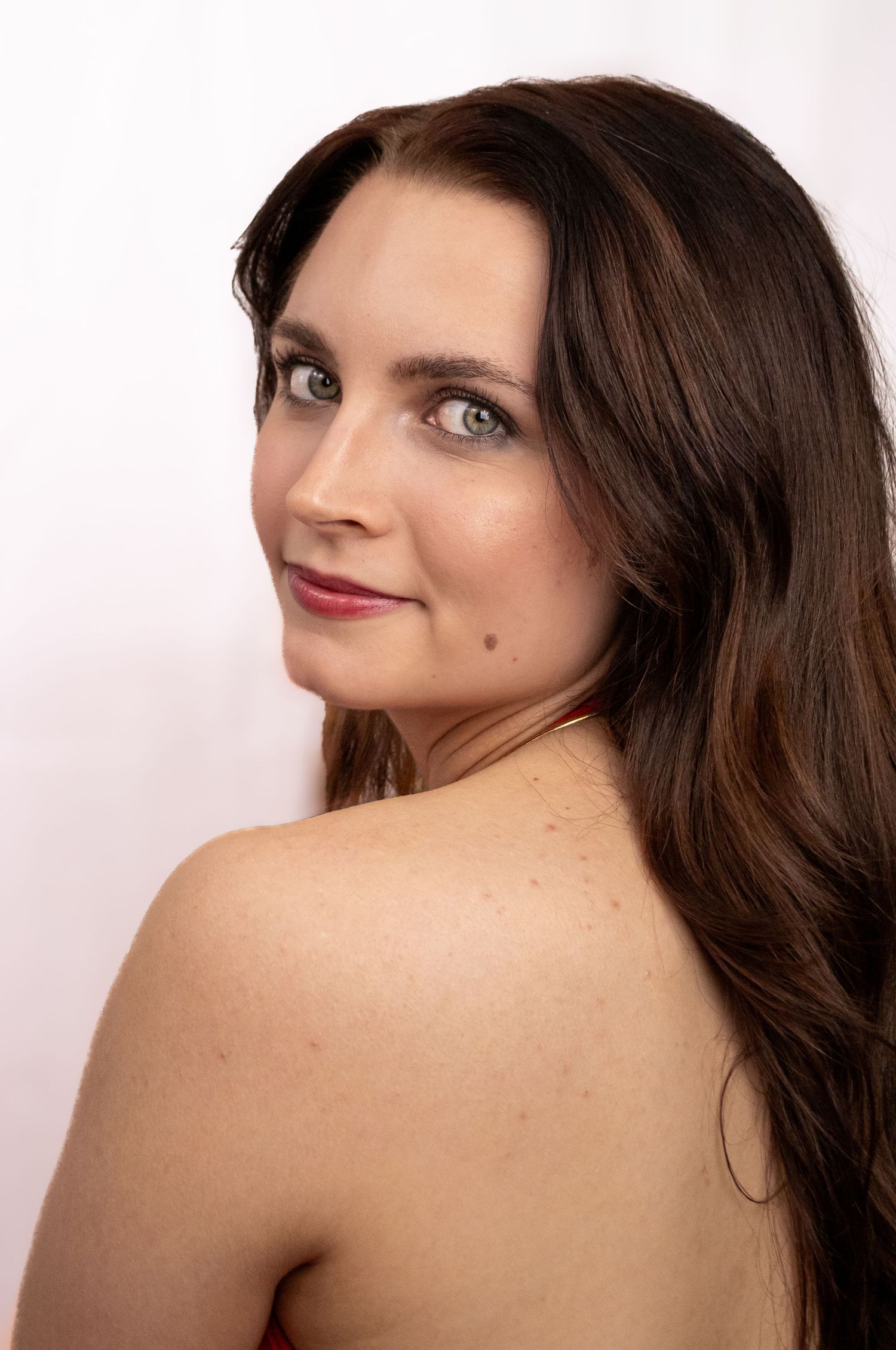 Portrait of a woman with long brown hair and red lipstick looking over her bare shoulder against a white background.