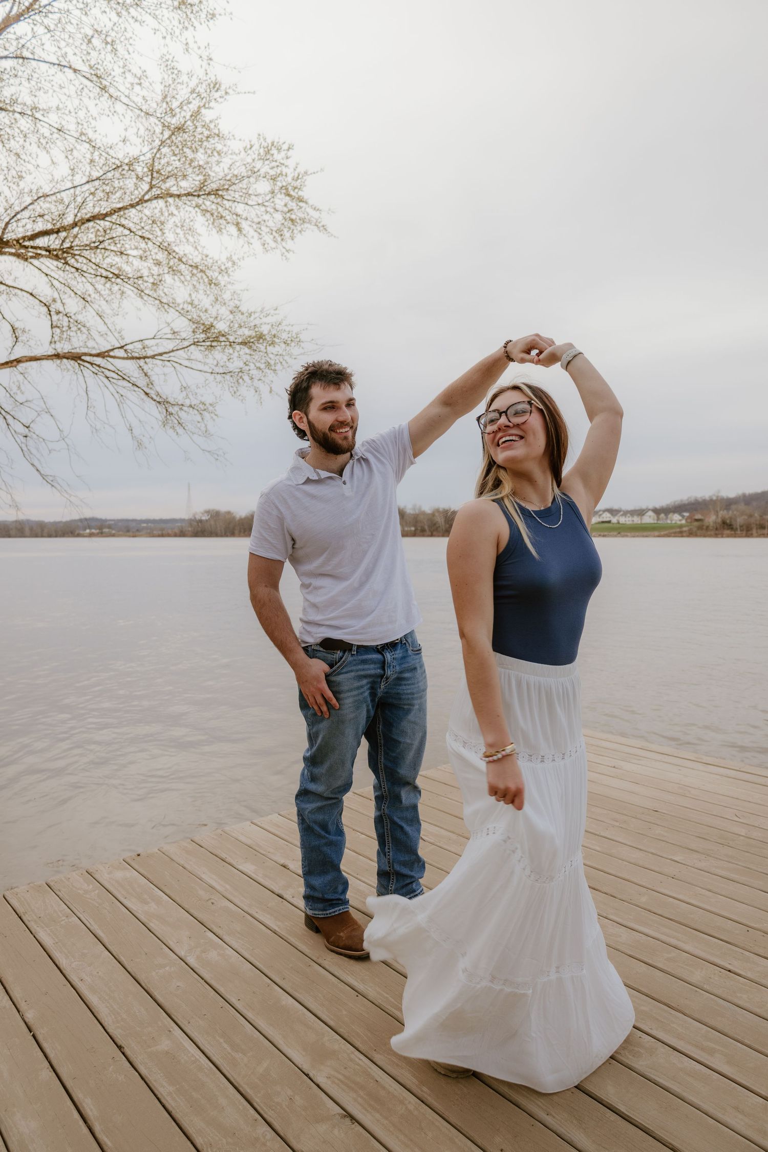 A couple shares a playful moment while dancing together on a wooden dock overlooking a serene lake.