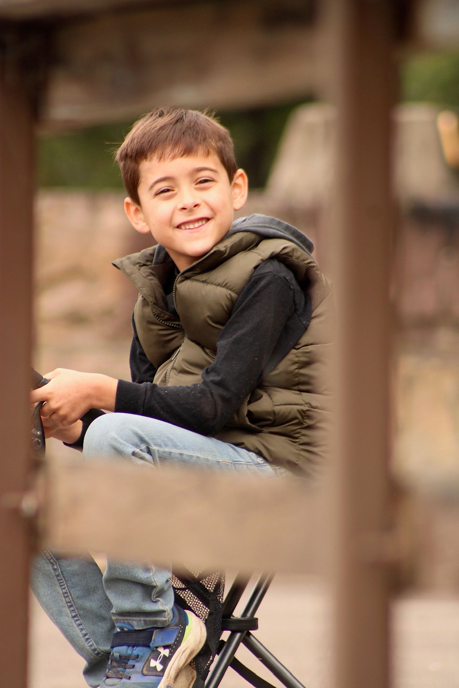 A child in a green jacket and jeans smiles while playing on outdoor playground equipment.