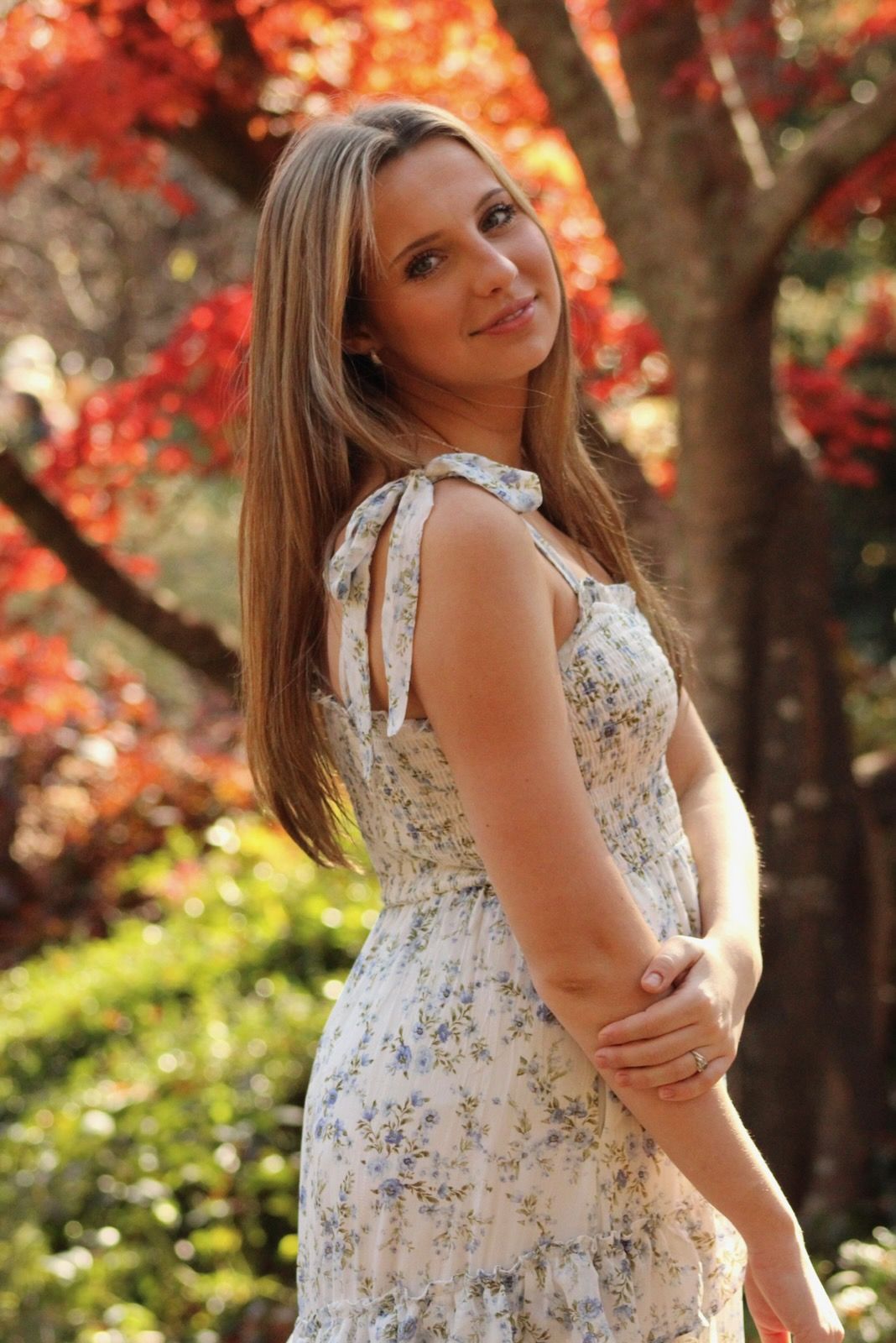 A person in a floral dress poses against vibrant red autumn foliage in a garden setting.