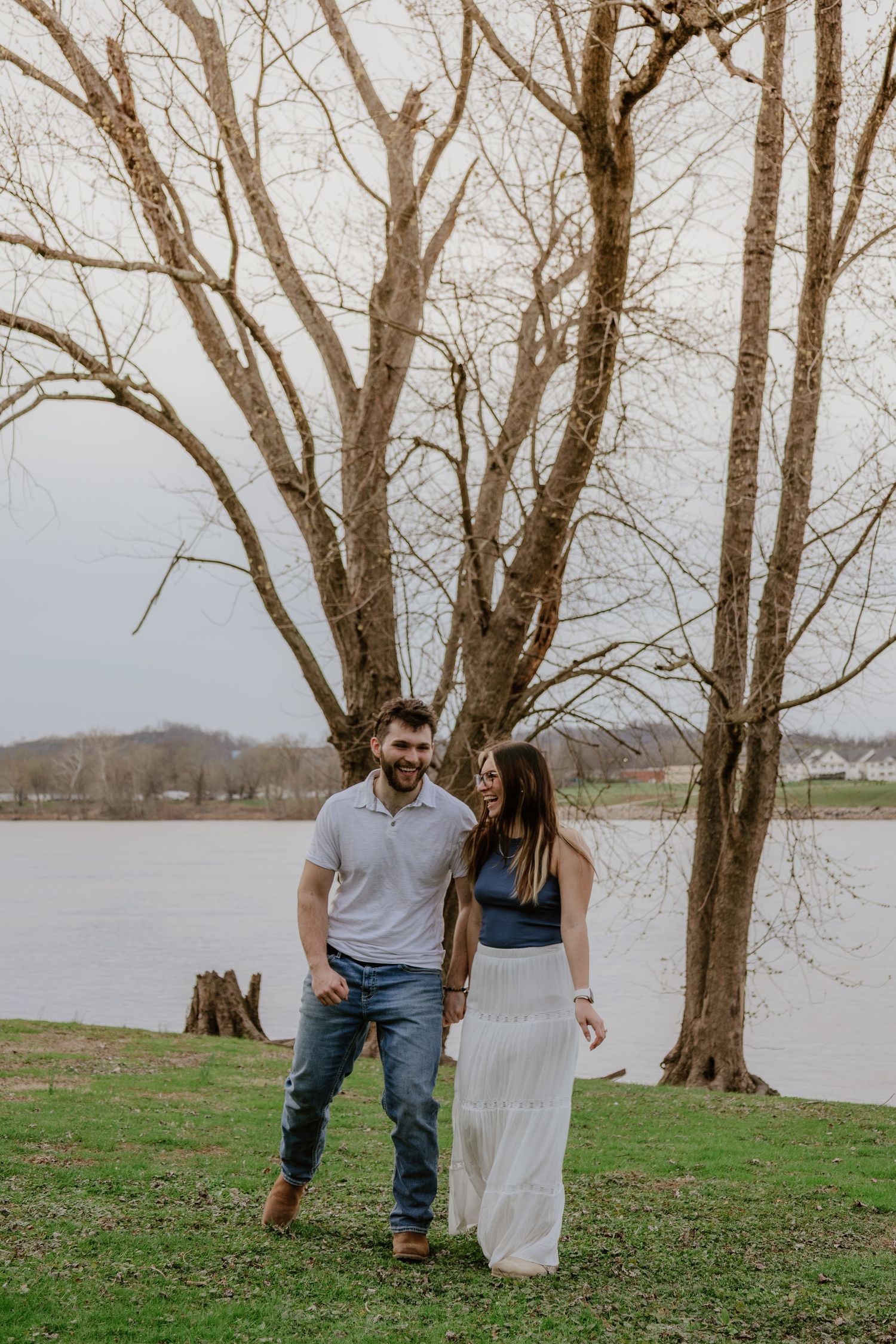 A couple walks together on a grassy shoreline with bare trees and a lake in the background on an overcast day.