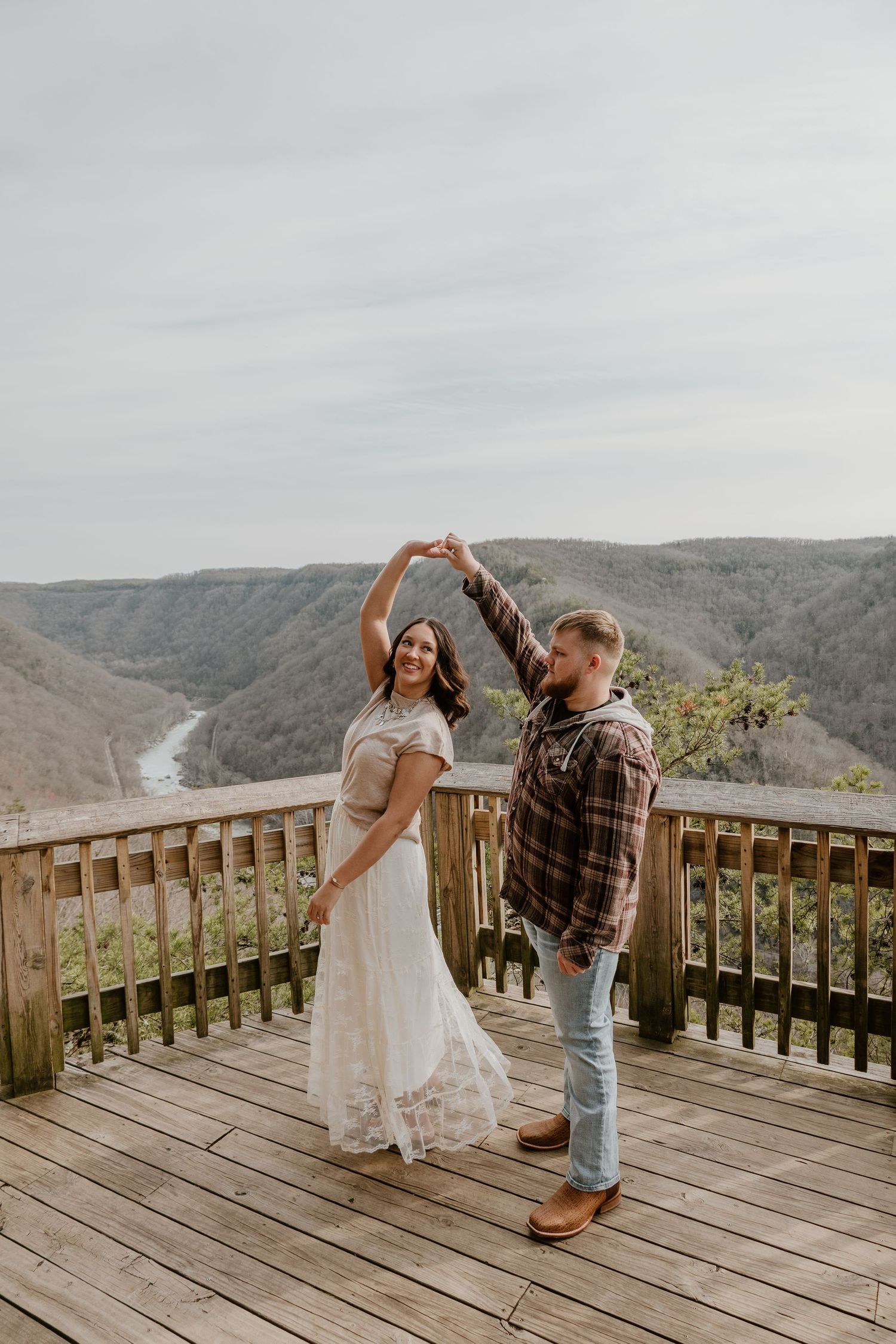 A couple dances together on a wooden observation deck overlooking a scenic mountain valley on a cloudy day.