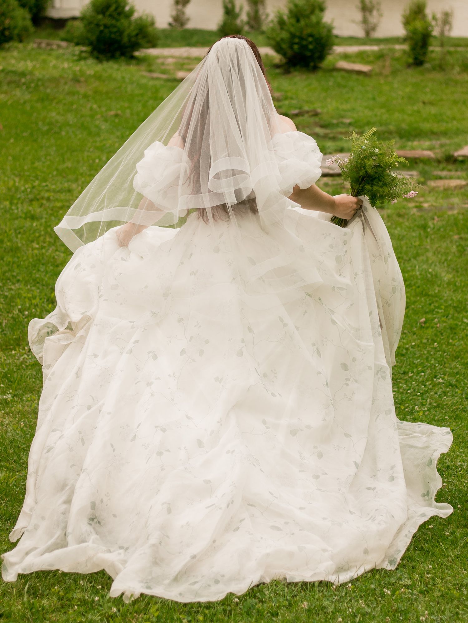 A flowing white wedding dress and veil photographed from behind on a grassy lawn.
