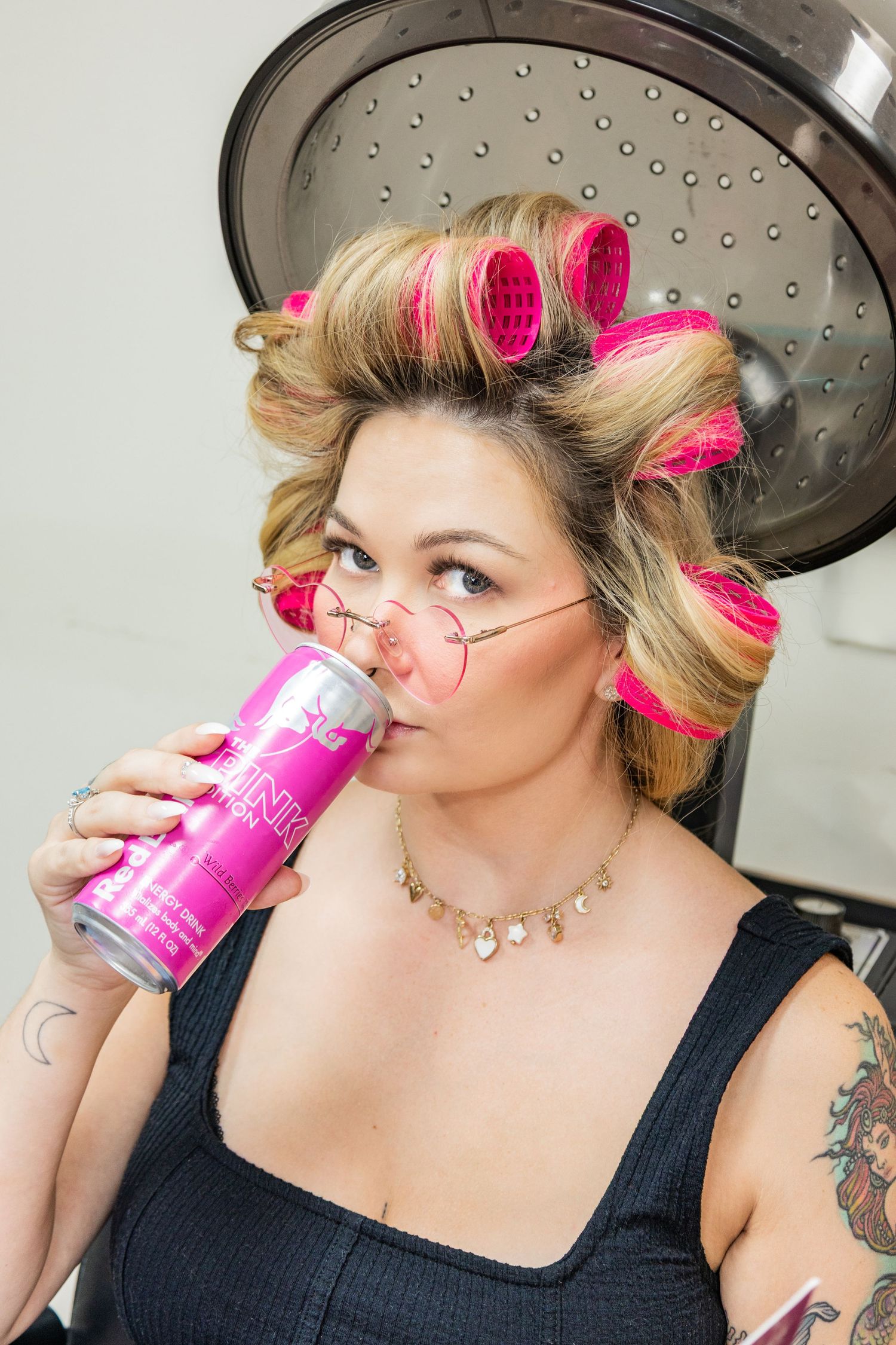 Person with pink hair rollers sips from a pink beverage can while getting ready in front of a salon mirror.