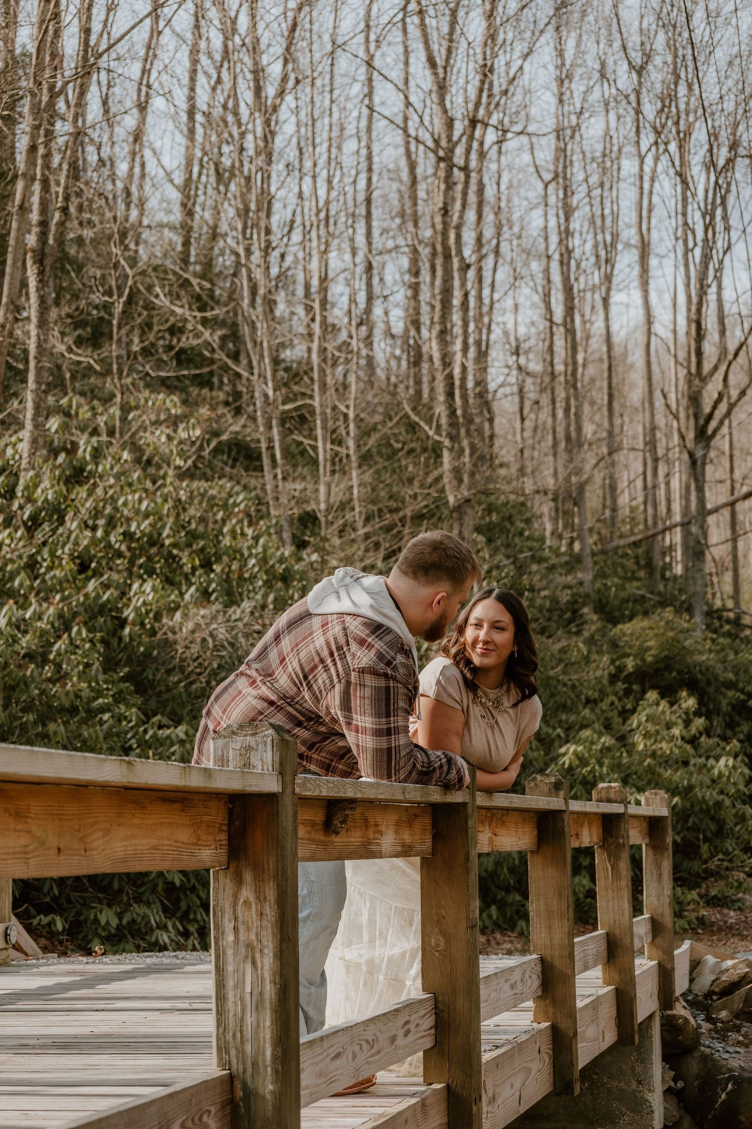 A couple shares a romantic moment on a wooden bridge surrounded by bare winter trees and evergreens.