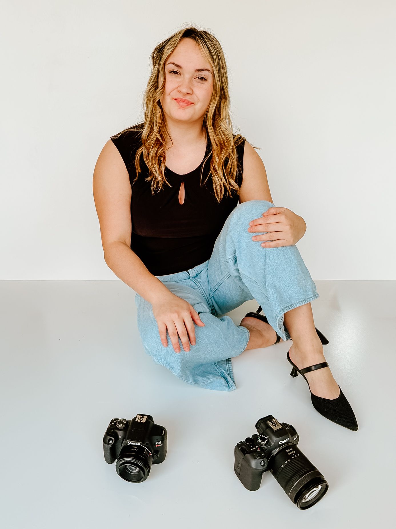 A photographer in black top and light denim sits casually with professional cameras on a white studio floor.