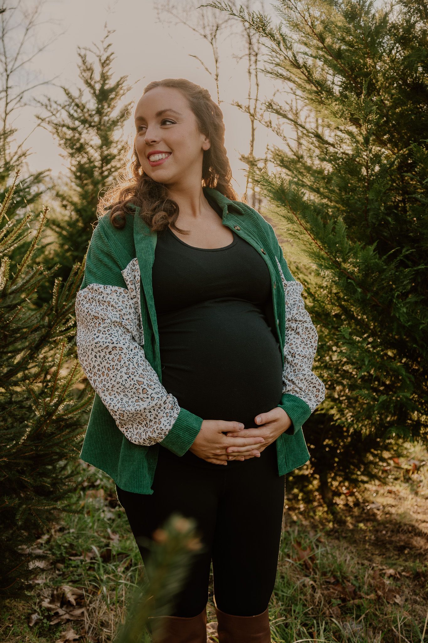 A person wearing a green cardigan and polka dot sleeves poses outdoors against evergreen trees at sunset.
