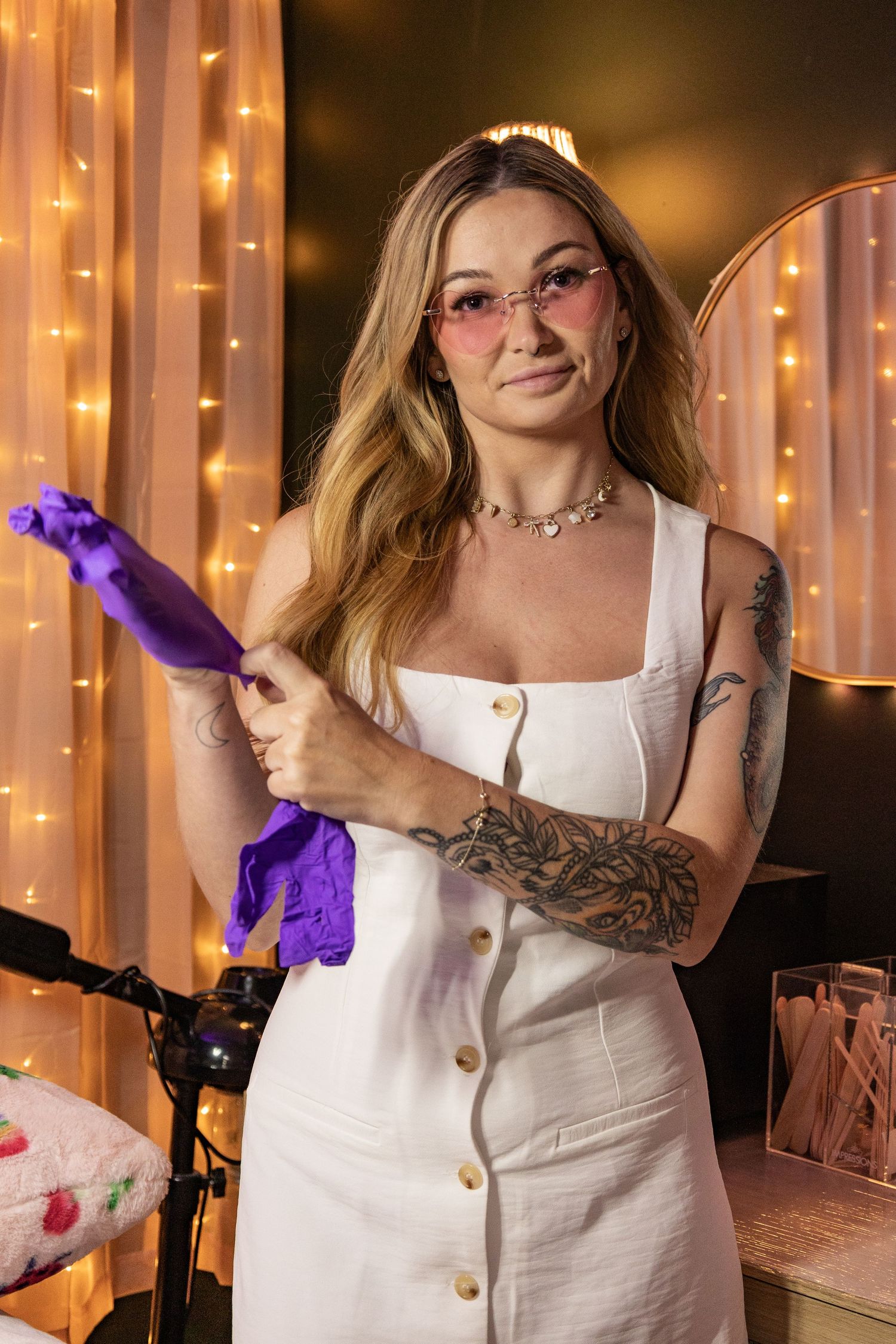 Woman in white dress holds purple objects while posing against a backdrop of glowing string lights.