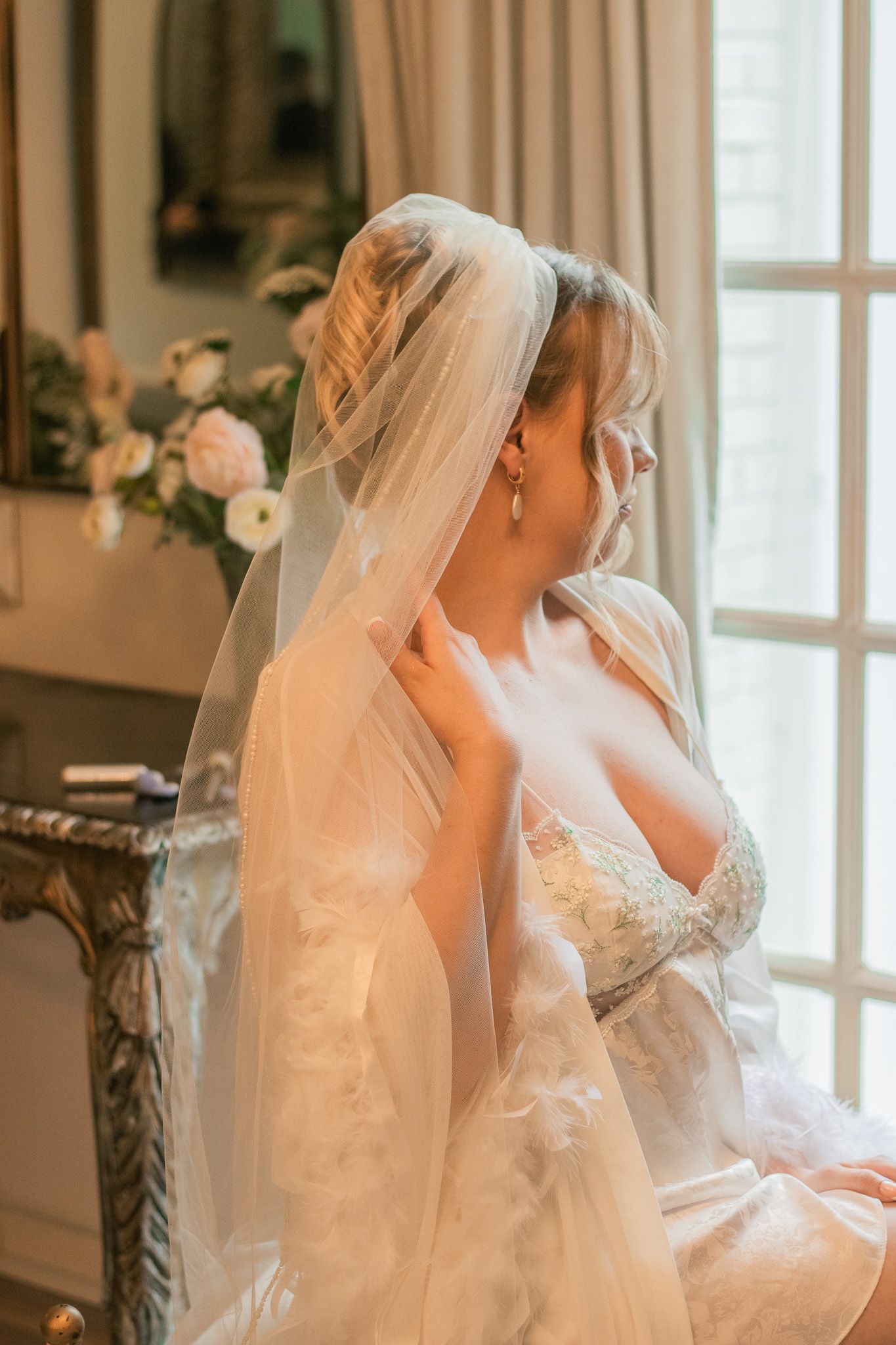 A bride in an elegant lace wedding dress and flowing veil sits by a window with soft natural lighting and floral decor.