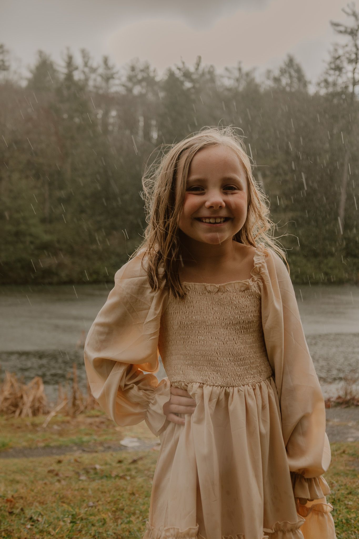 A young child in a beige smocked dress smiles outdoors on a cloudy day near a lake.