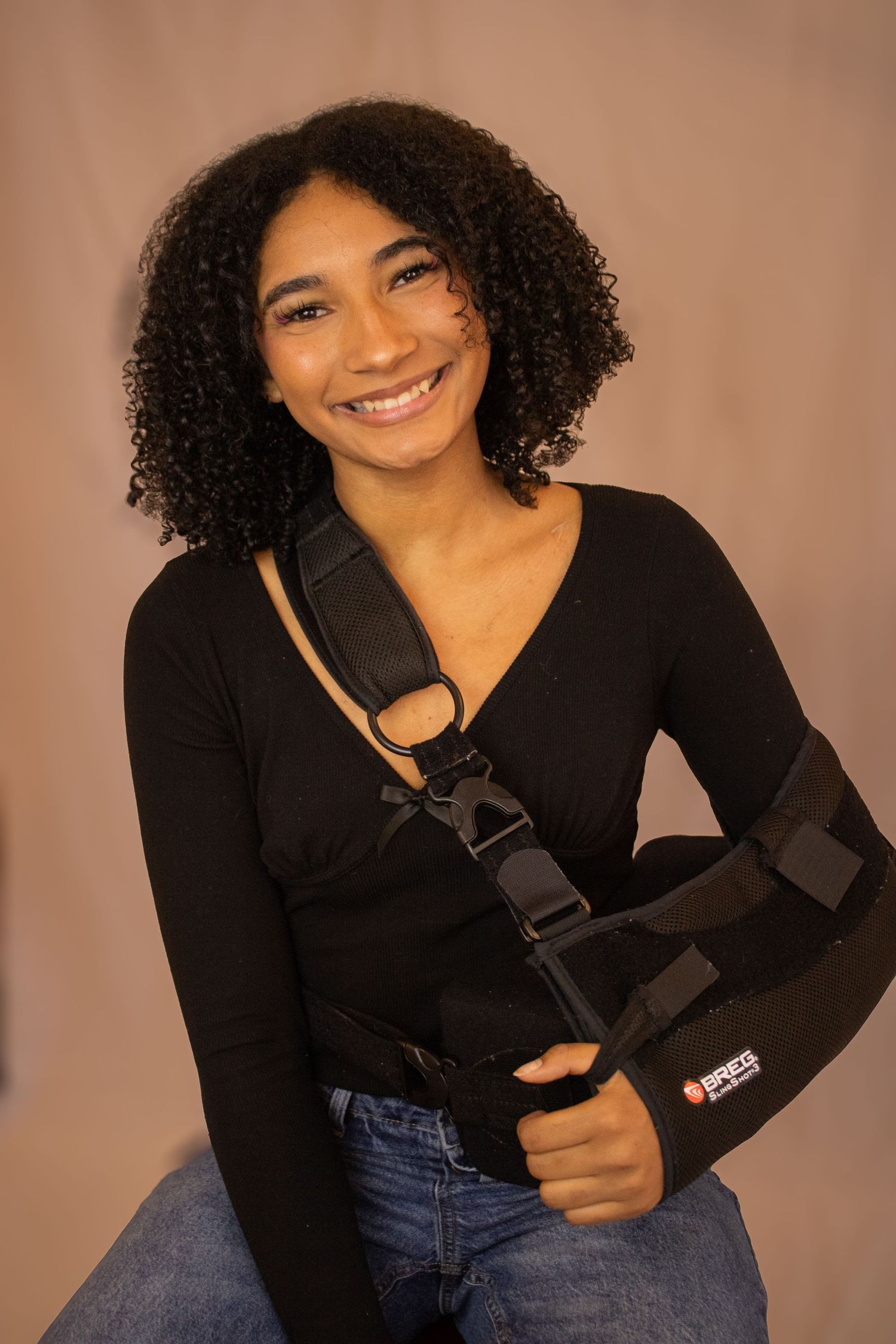 A person in a black sweater and jeans poses against a beige background while holding a camera strap.