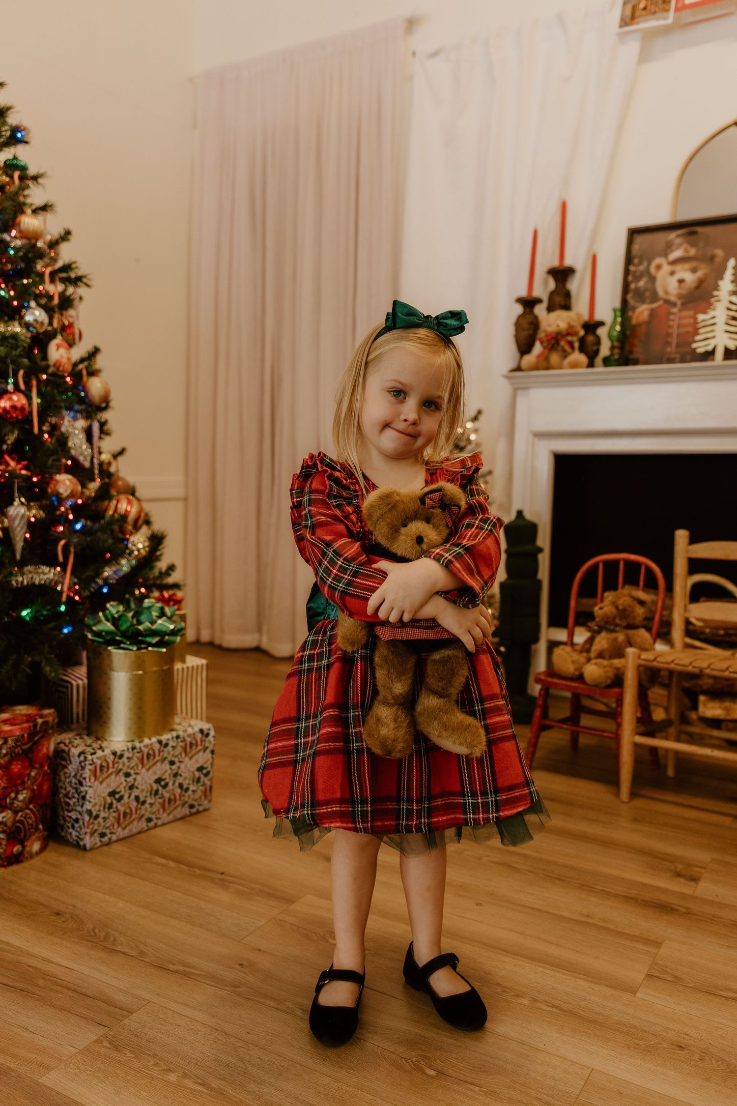 A child in a red plaid dress sits and plays near a decorated Christmas tree and wrapped presents in a cozy living room.