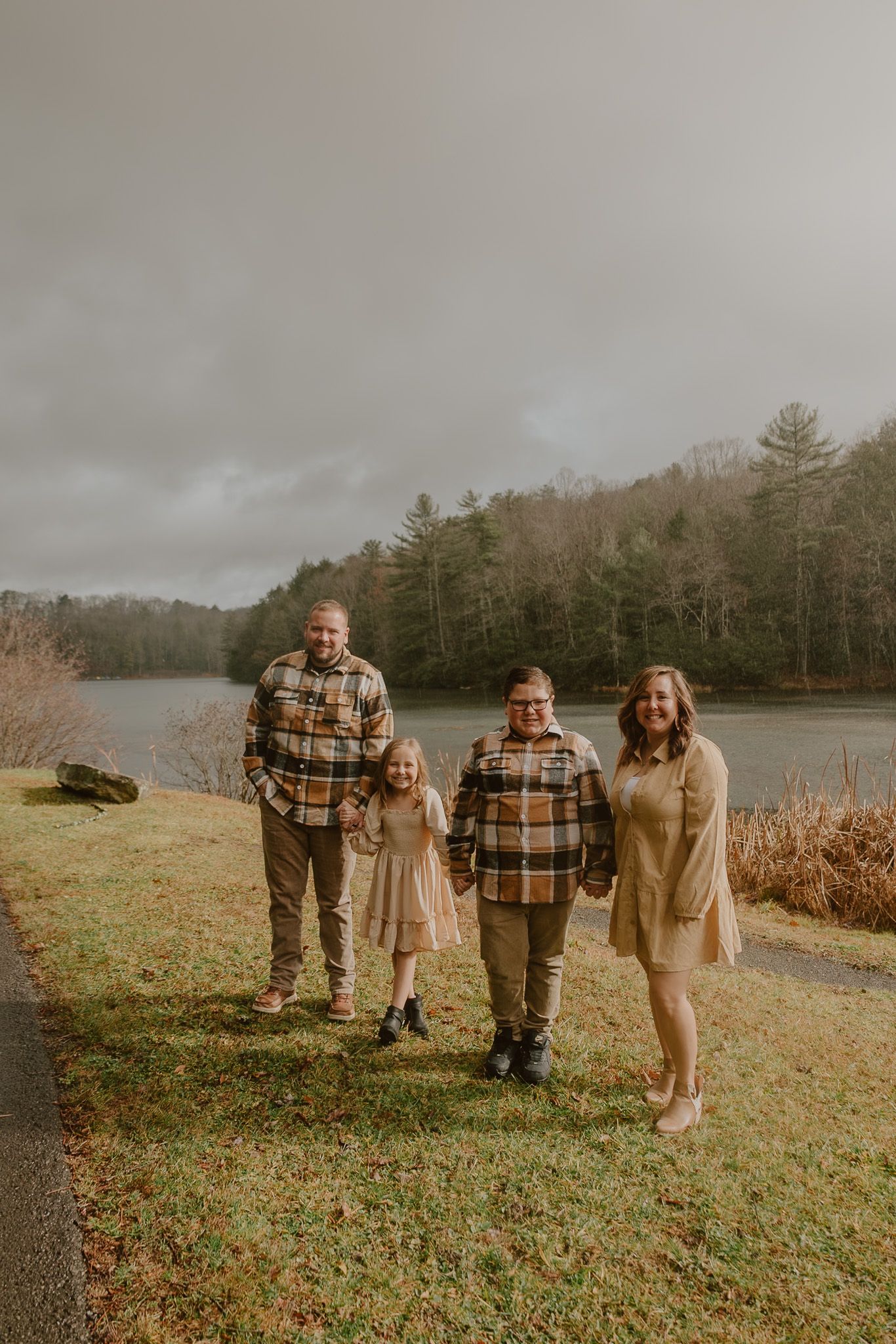 A family wearing coordinated plaid and beige outfits poses together on a grassy hill near a lake on an overcast day.