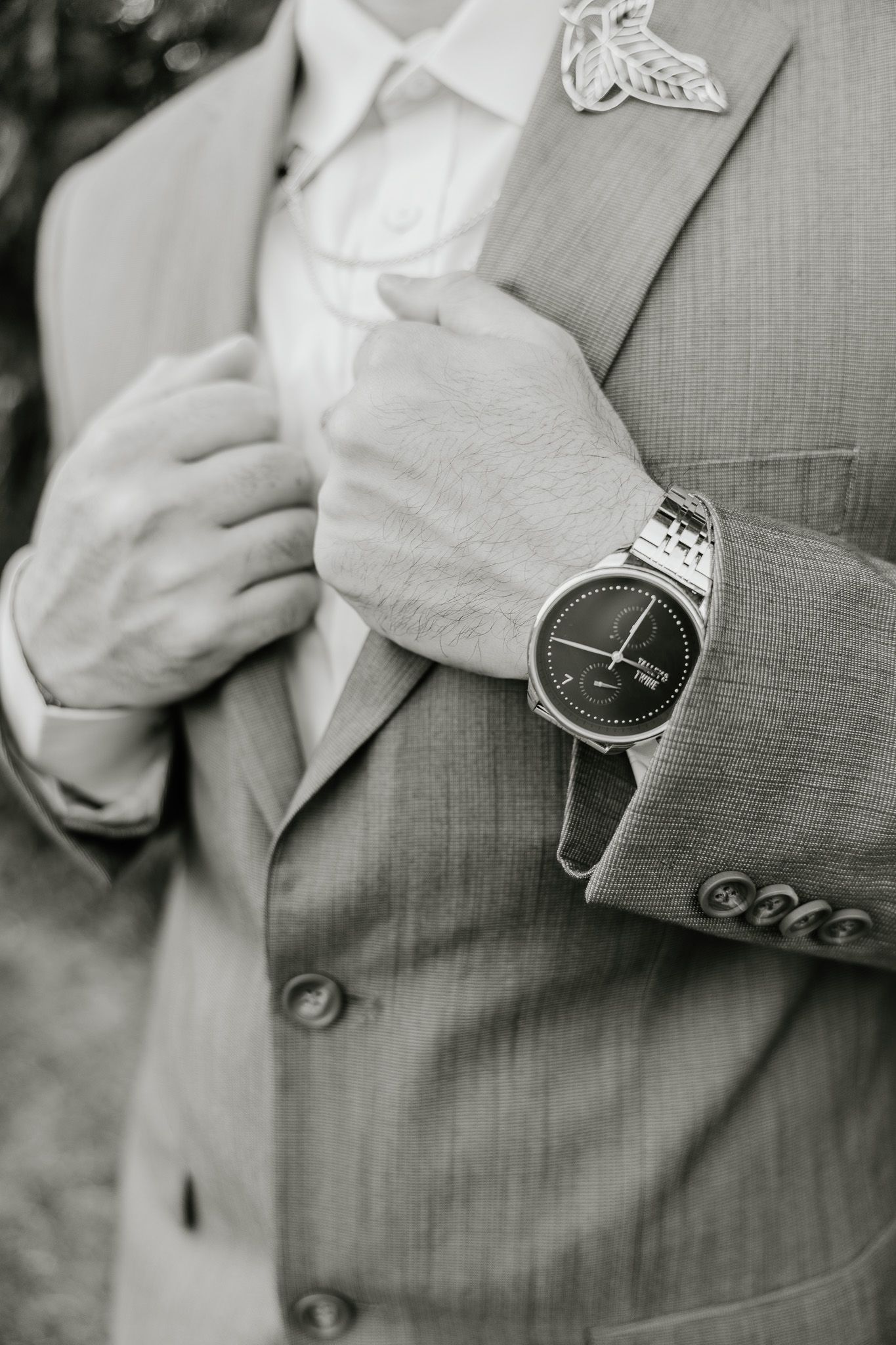 Close-up black and white photo of a suit jacket and wristwatch, showing formal wedding attire details.