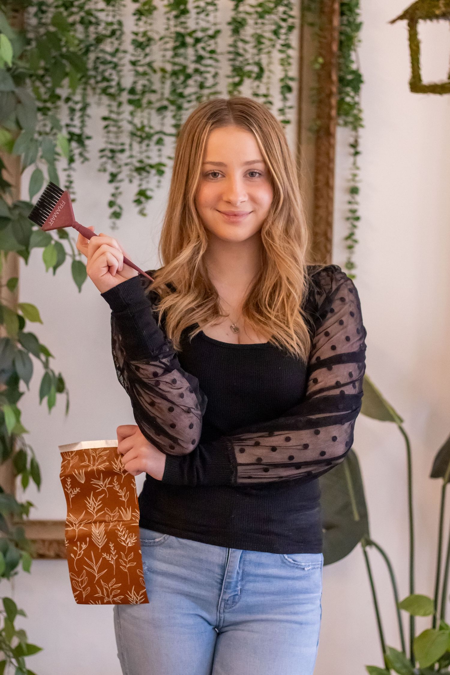 A person in a black polka dot sleeve top and jeans holds a brown gift bag in front of hanging plants and white wall decor.