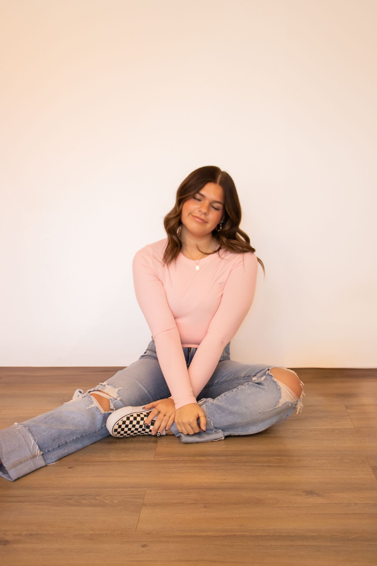 Person in pink sweater and ripped jeans relaxes on wooden floor in casual poses against light background.