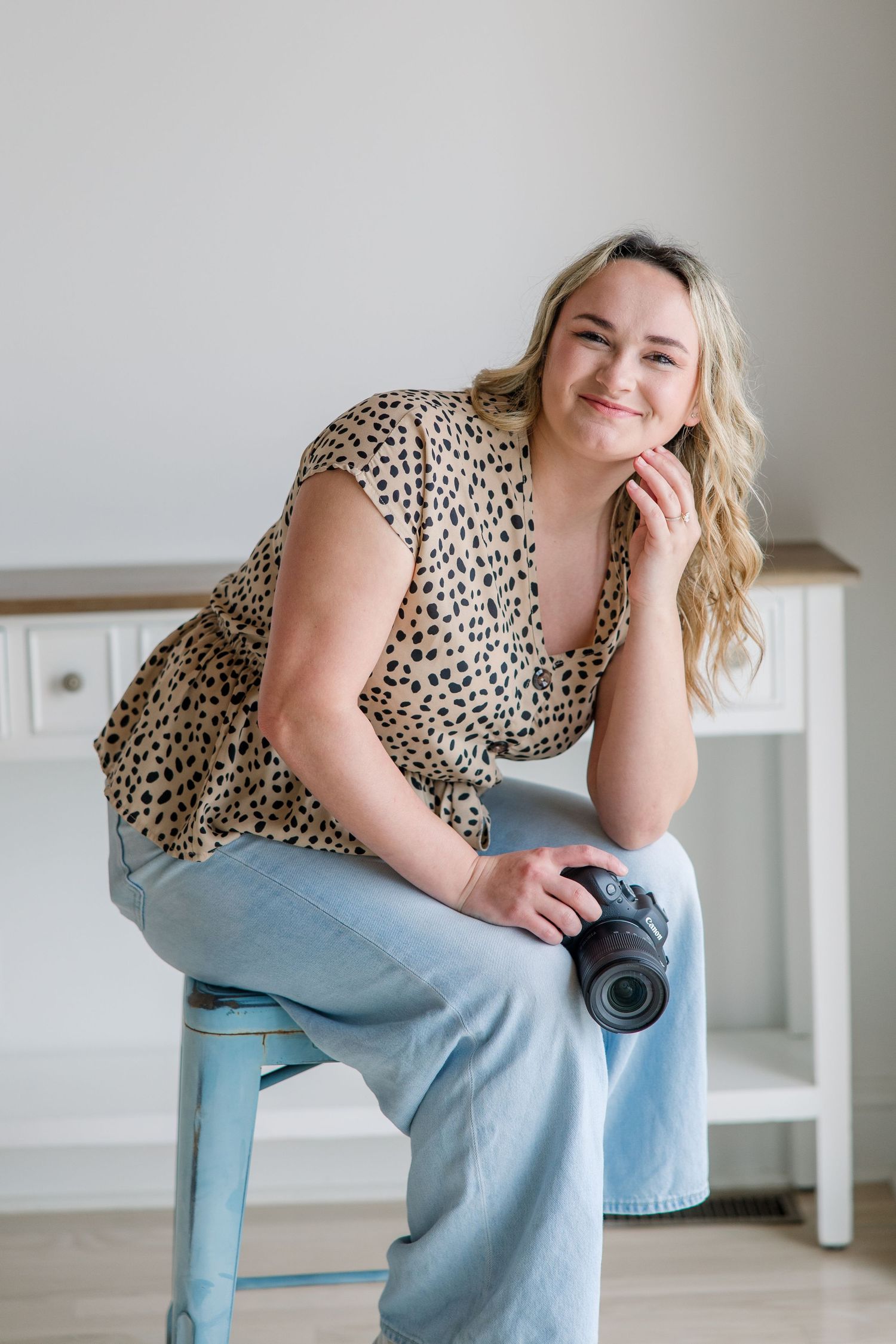 Casual portrait of someone in a leopard print top and jeans sitting on a blue stool against a white wall.