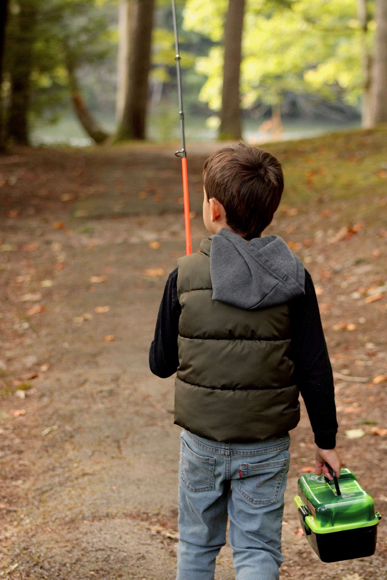 Child in puffy vest carries fishing rod and tackle box while walking on autumn forest trail.