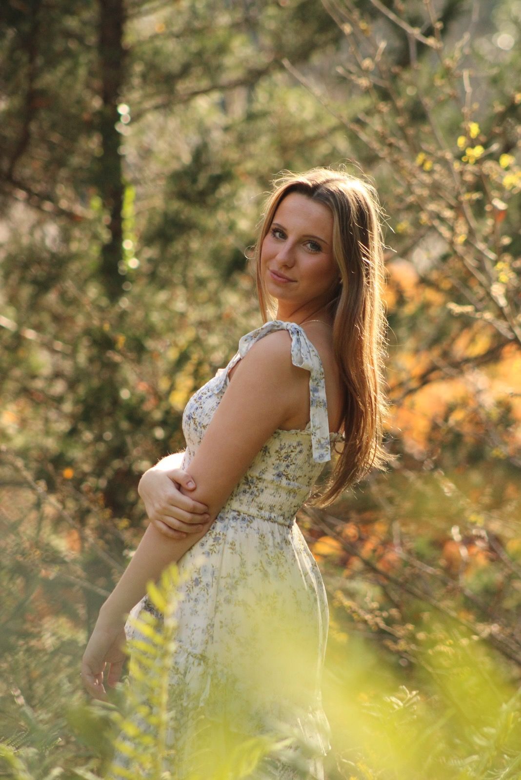 A woman in a white lace dress poses gracefully in a sunlit forest setting during a golden hour photoshoot.