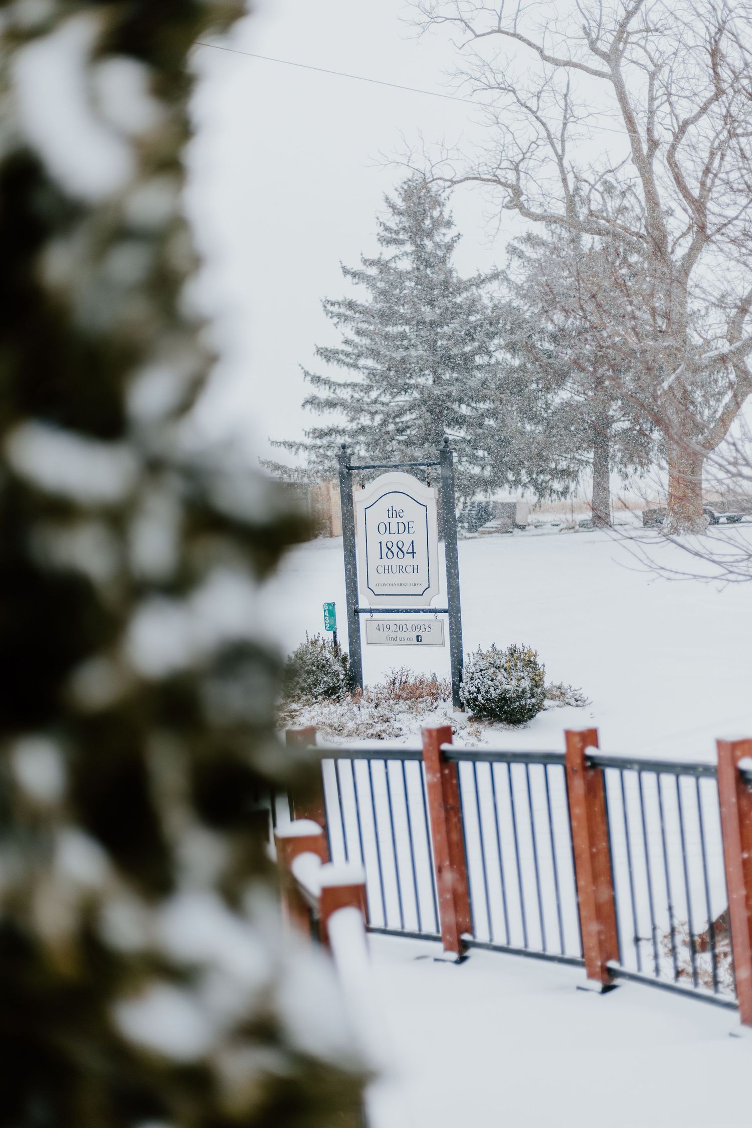A snowy winter scene with a white wooden sign post and railing covered in fresh snowfall against an evergreen tree backdrop.