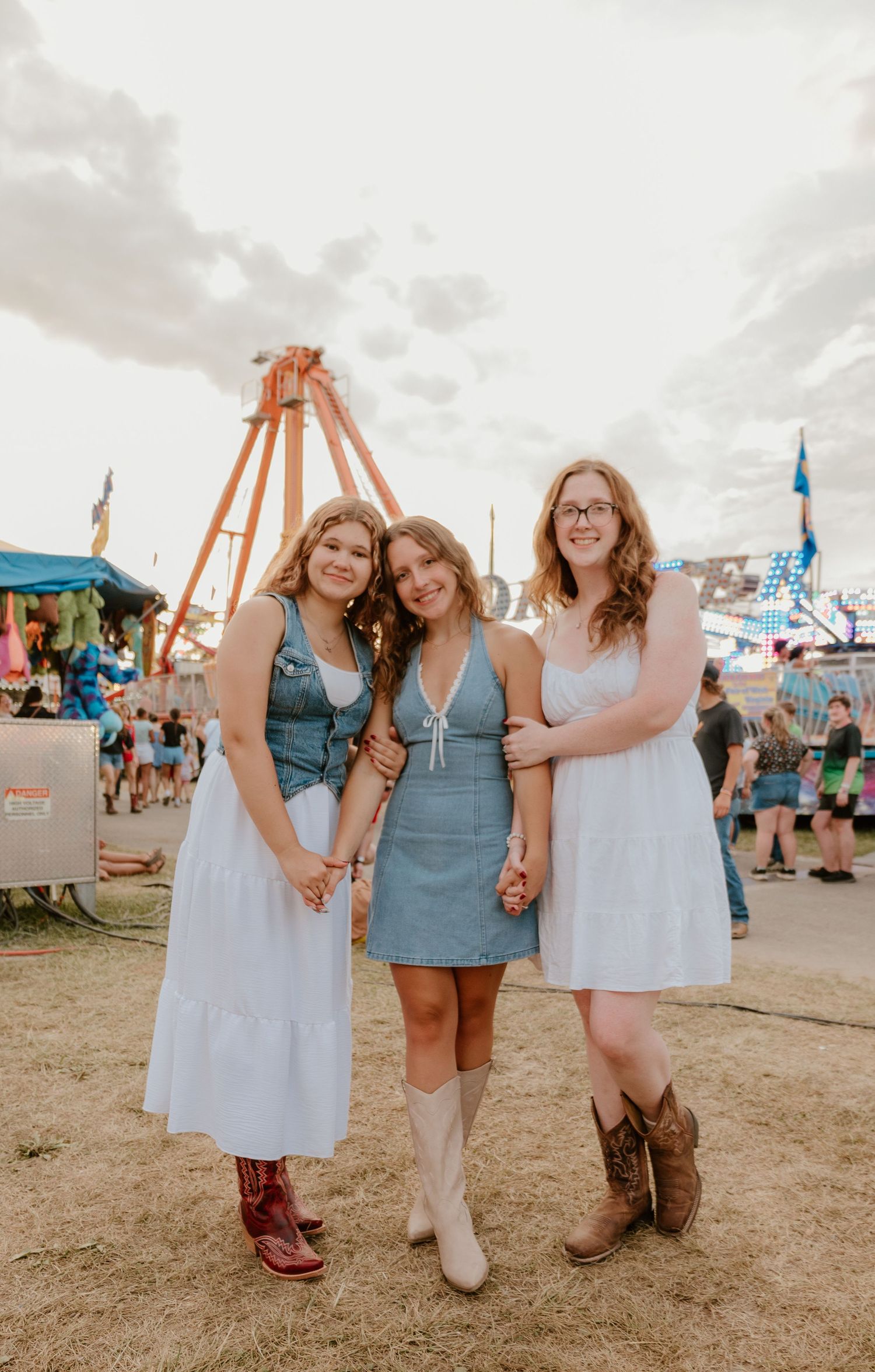 Three friends in denim and white dresses pose together at a county fair with carnival rides in the background.